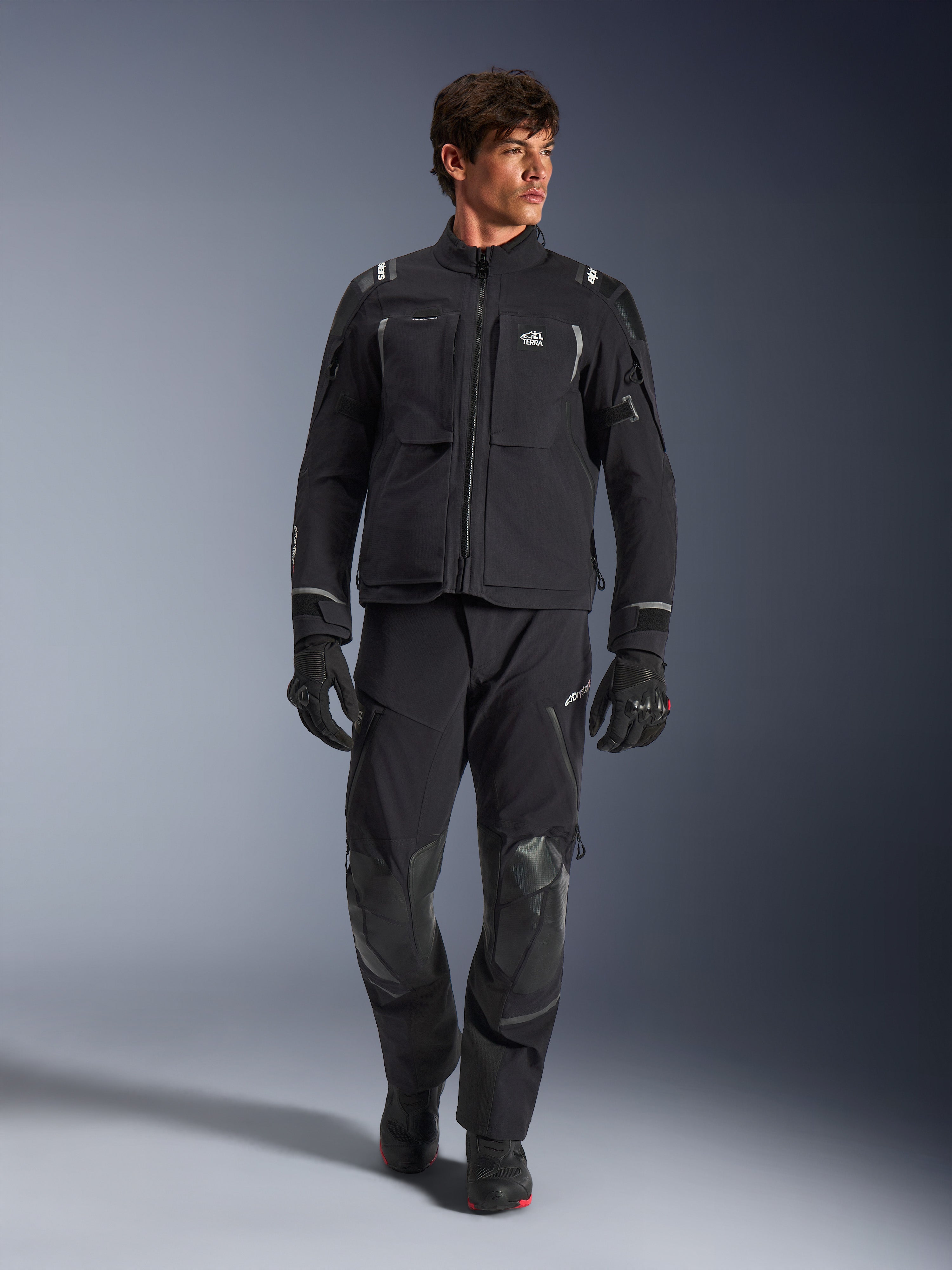 Modelo masculino a usar o Casaco Alpinestars Andes Pro Drystar® XF, casaco de motociclismo de aventura em preto e cinzento escuro, combinado com calças técnicas e luvas a condizer, com múltiplos bolsos e a marca 'Terra' no peito.