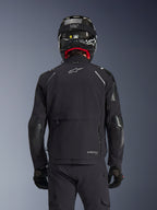 Casaco Alpinestars Andes Pro Drystar® XF em preto, vista traseira de um motociclista com capacete de fibra de carbono, compartimento de armazenamento traseiro de grandes dimensões e logótipo Alpinestars branco, equipamento técnico de aventura para todas as estações