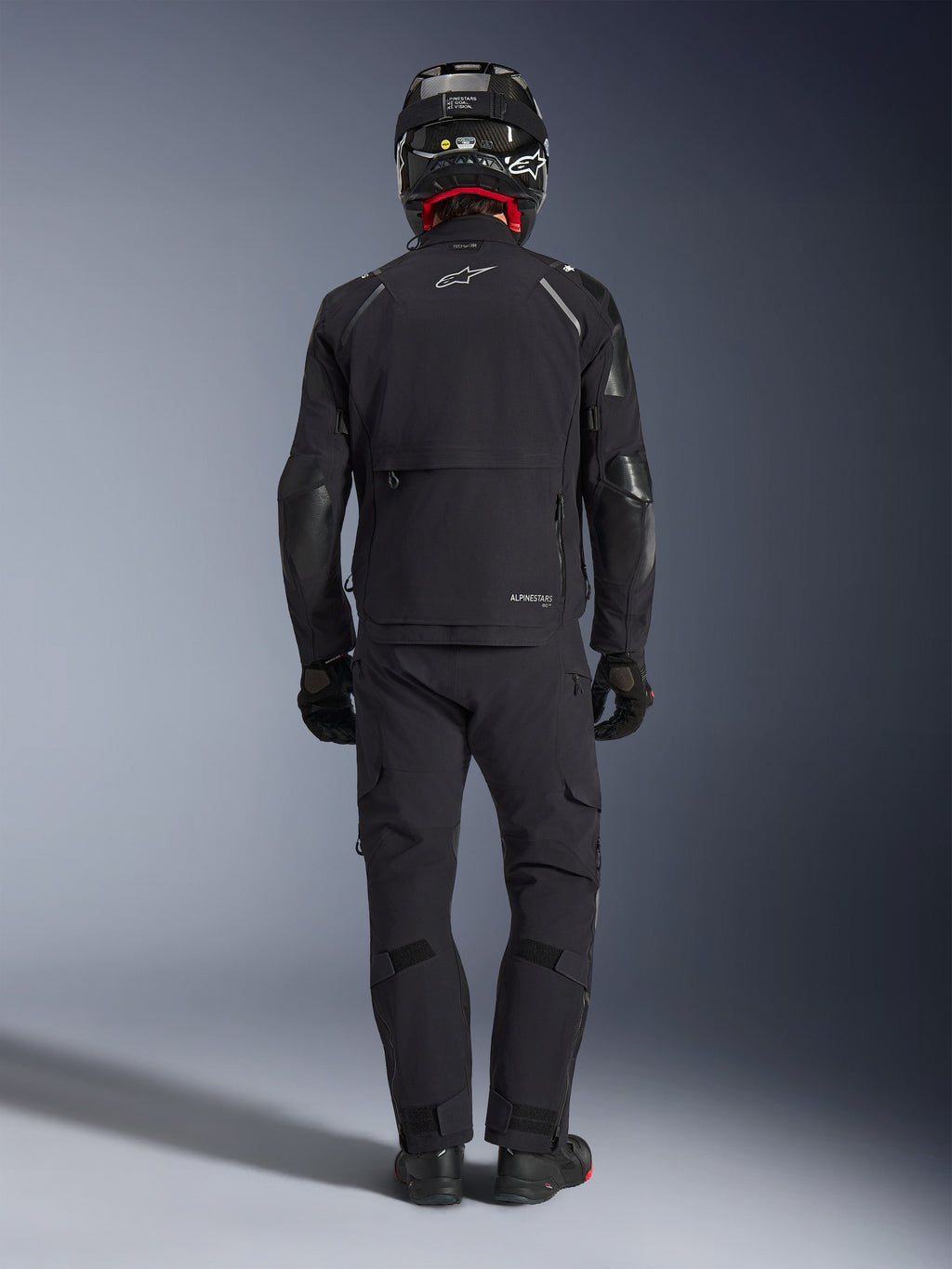Casaco Alpinestars Andes Pro Drystar® XF em preto e cinzento escuro, vista traseira de um motociclista com equipamento completo de adventure touring, incluindo calças a condizer e capacete de fibra de carbono, apresentando um grande bolso de carga traseiro e logótipo refletor da Alpinestars nas costas.