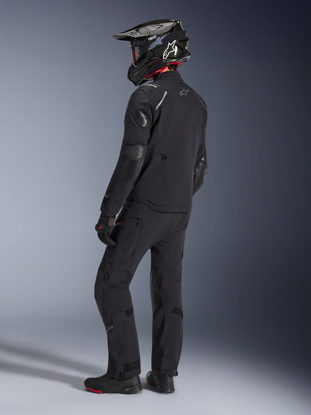 Motociclista a usar o Casaco Alpinestars Andes Pro Drystar® XF e calças a condizer em Preto e Cinzento Escuro, vista de perfil traseiro, com capacete de aventura em fibra de carbono preto, luvas pretas e botas de aventura, equipamento coordenado de alta performance para touring de aventura.
