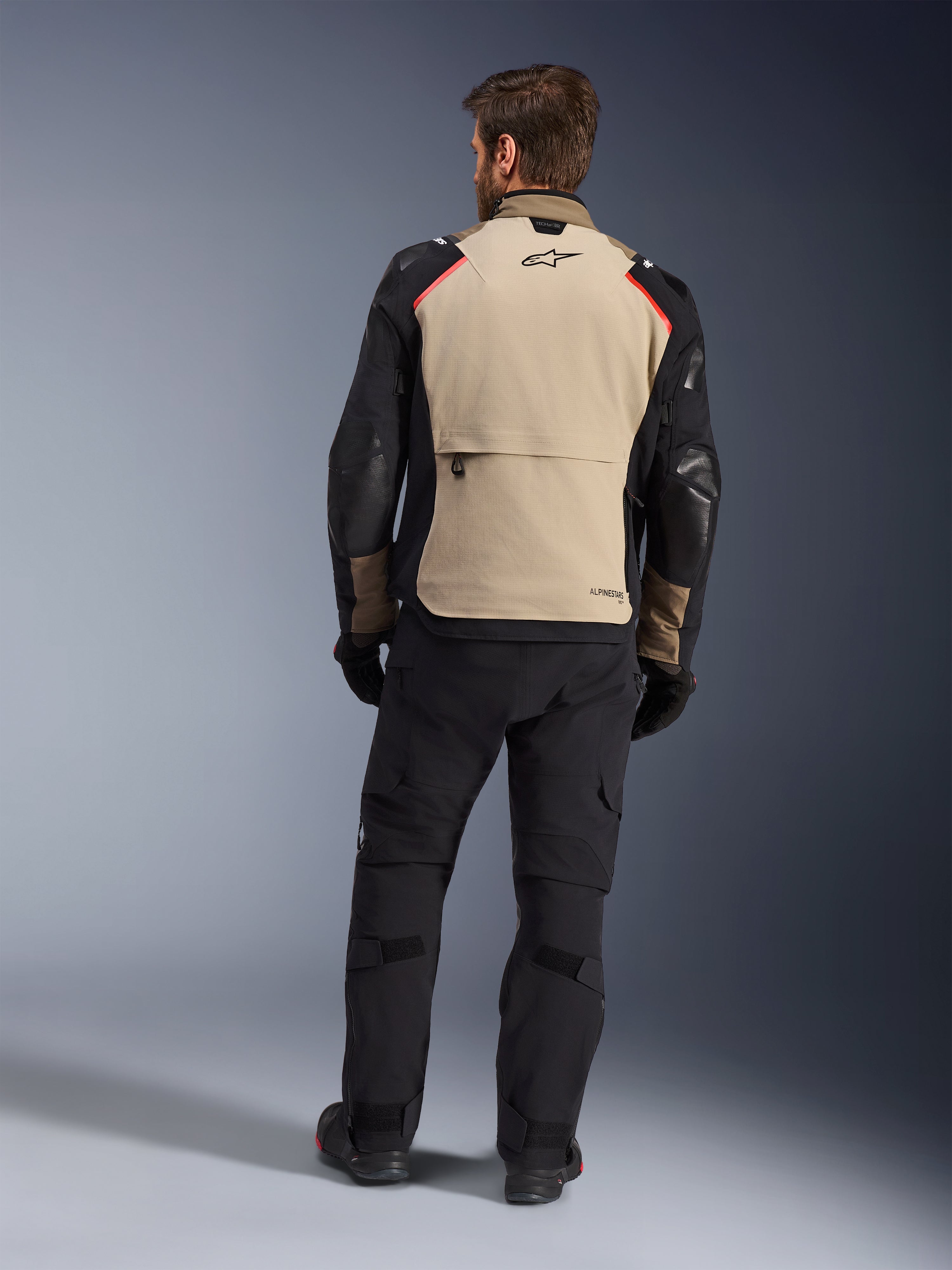 Casaco Alpinestars Andes Pro Drystar® XF, Walnut Aluminum, vista traseira, casaco de aventura para motociclista com mangas pretas e detalhes vermelhos nos ombros, usado com calças de condução e luvas pretas.