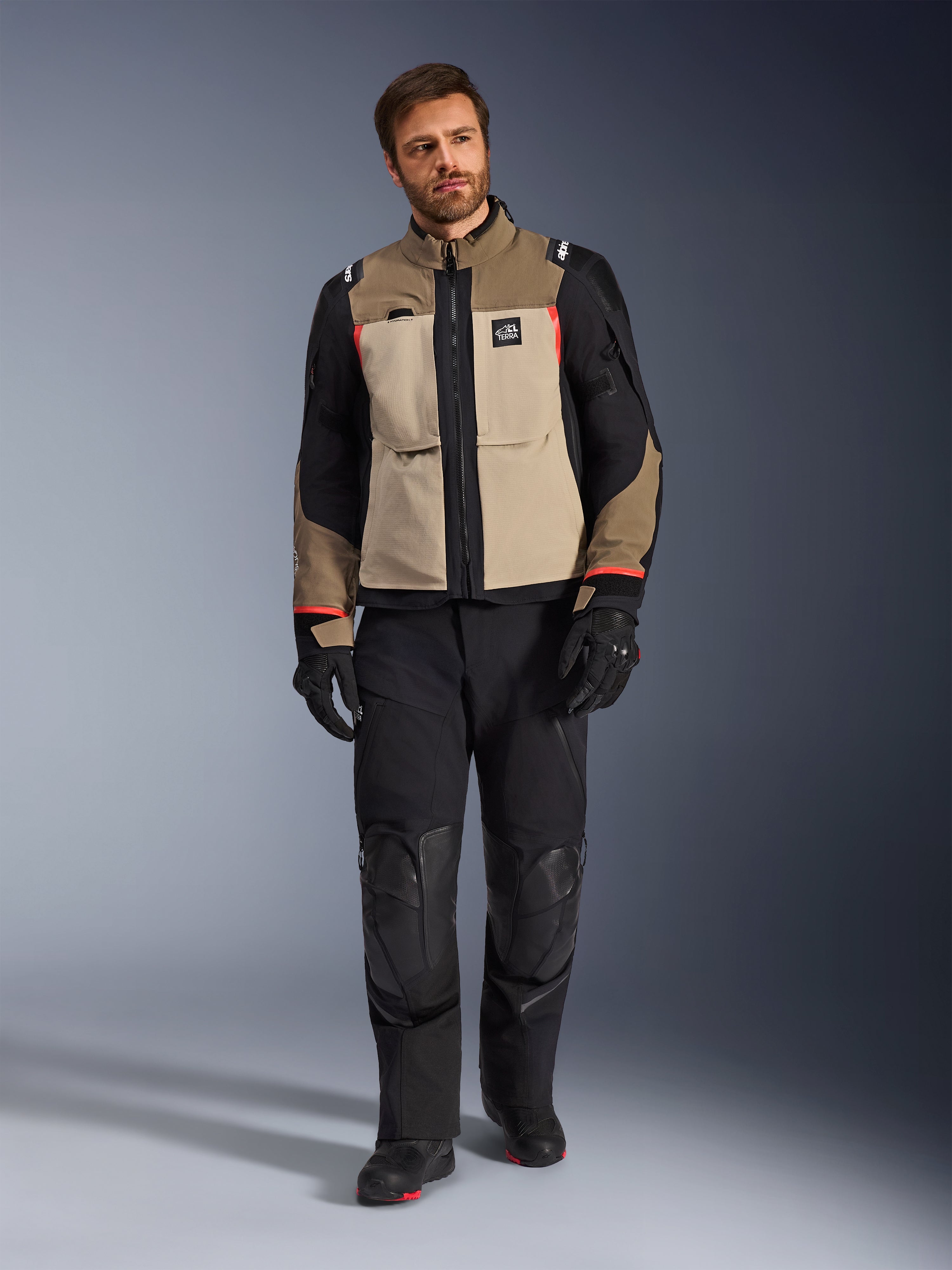 Modelo masculino a usar o casaco Alpinestars Andes Pro Drystar® XF em Walnut Aluminum, casaco de aventura e turismo bege e preto com detalhes em vermelho, combinado com calças, luvas e botas de motociclismo pretas, fotografia de corpo inteiro em estúdio de equipamento de proteção para condução.