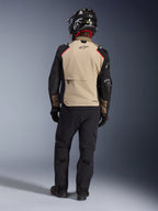 Piloto visto de costas a usar o casaco Alpinestars Andes Pro Drystar® XF em Walnut Aluminum, painel traseiro bege com mangas pretas e detalhes a vermelho, combinado com calças técnicas de aventura pretas e capacete de motociclista, equipamento de touring premium para todas as condições meteorológicas.