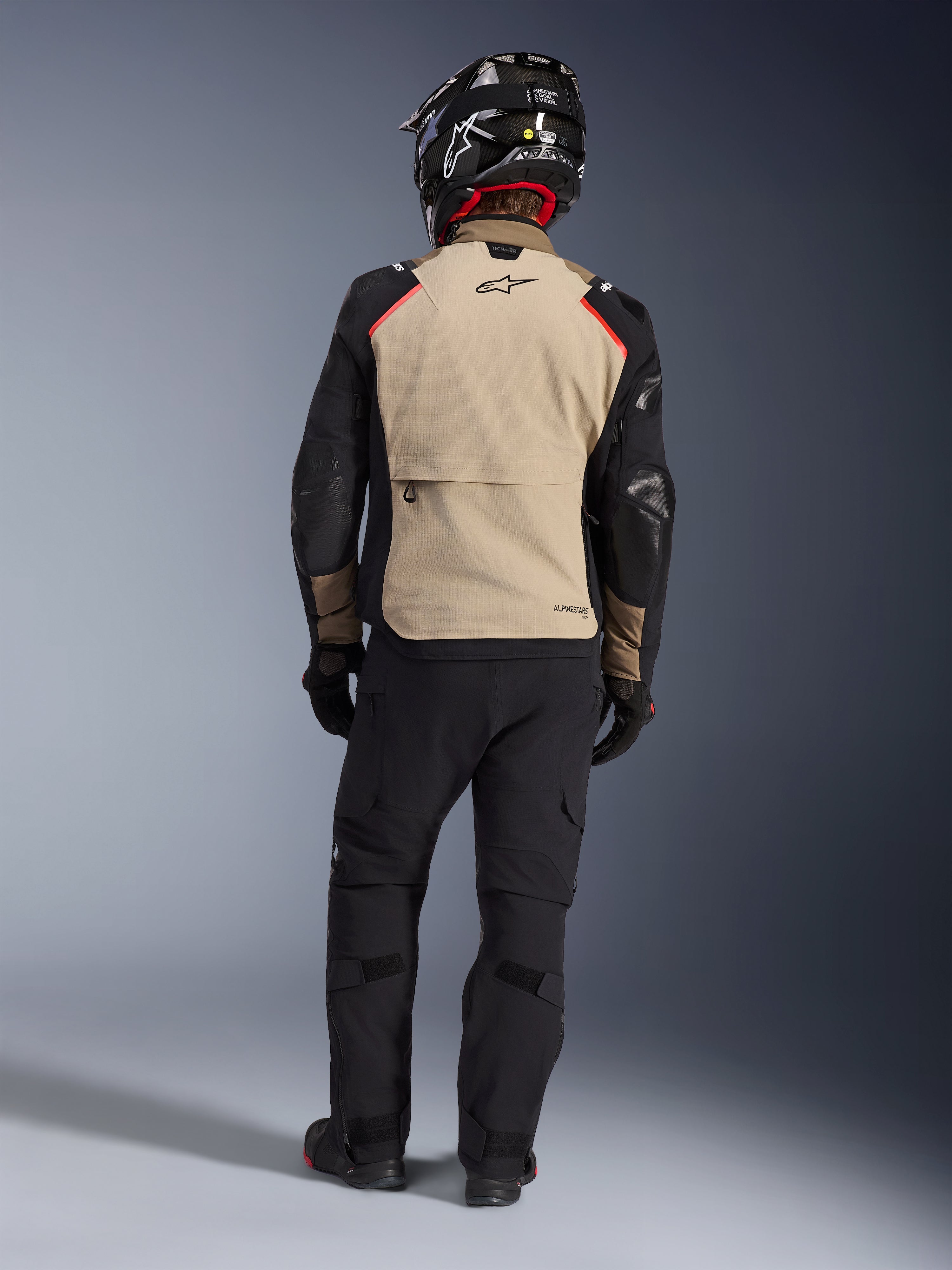 Piloto visto de costas a usar o casaco Alpinestars Andes Pro Drystar® XF em Walnut Aluminum, painel traseiro bege com mangas pretas e detalhes a vermelho, combinado com calças técnicas de aventura pretas e capacete de motociclista, equipamento de touring premium para todas as condições meteorológicas.