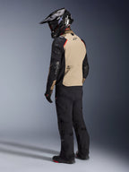 Motociclista visto de costas com o casaco Alpinestars Andes Pro Drystar® XF em Walnut Aluminum, com um painel traseiro bege, mangas pretas e detalhes em vermelho, capacete de aventura preto, calças de condução pretas e botas pretas, equipamento profissional de touring de aventura.