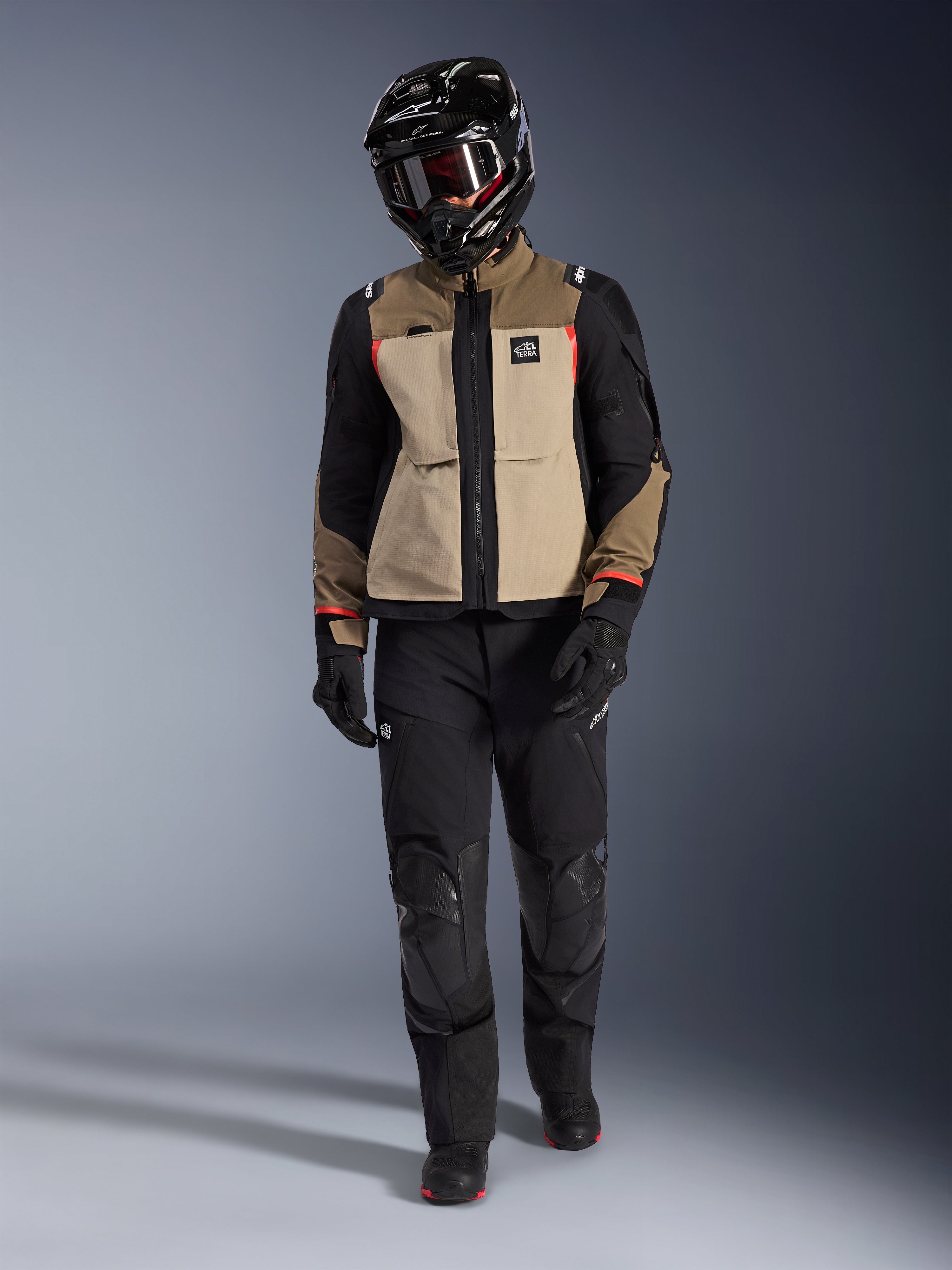 Casaco Alpinestars Andes Pro Drystar® XF em Walnut Aluminum, usado com calças de aventura pretas, luvas pretas e capacete integral preto com óculos, com detalhes em vermelho e marca All Terra, conjunto profissional de adventure touring