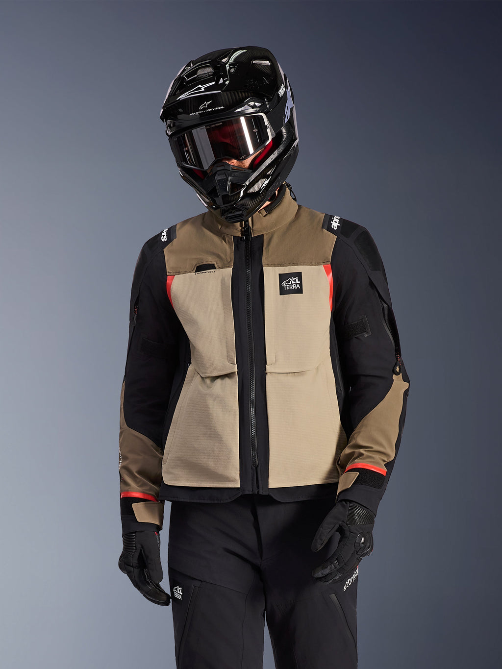 Casaco Alpinestars Andes Pro Drystar® XF, casaco de motociclismo para touring de aventura, em bege walnut aluminum e castanho com painéis pretos e pormenores vermelhos, usado com um capacete e óculos Alpinestars pretos, equipamento técnico de motociclismo