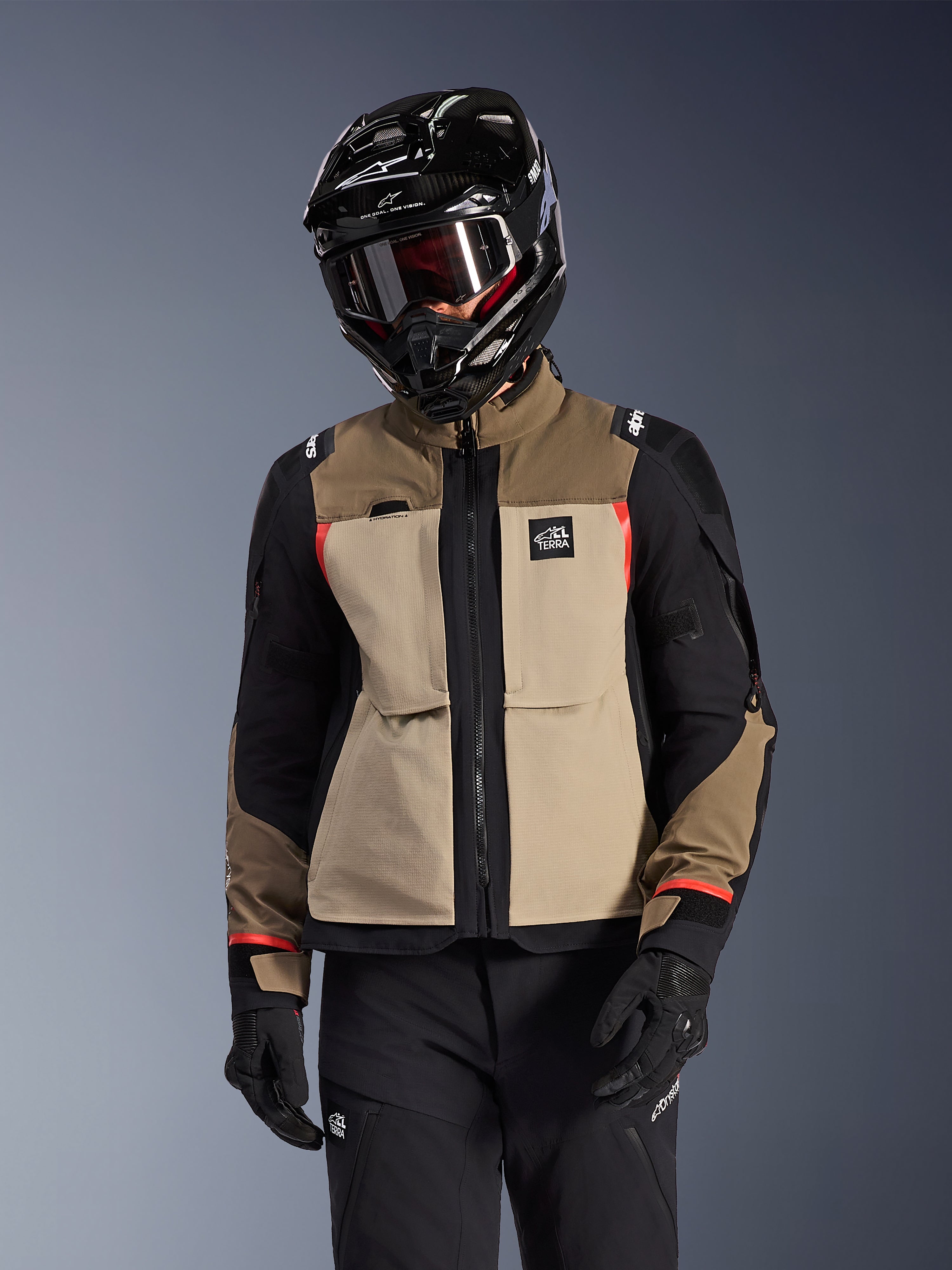 Casaco Alpinestars Andes Pro Drystar® XF, casaco de motociclismo para touring de aventura, em bege walnut aluminum e castanho com painéis pretos e pormenores vermelhos, usado com um capacete e óculos Alpinestars pretos, equipamento técnico de motociclismo