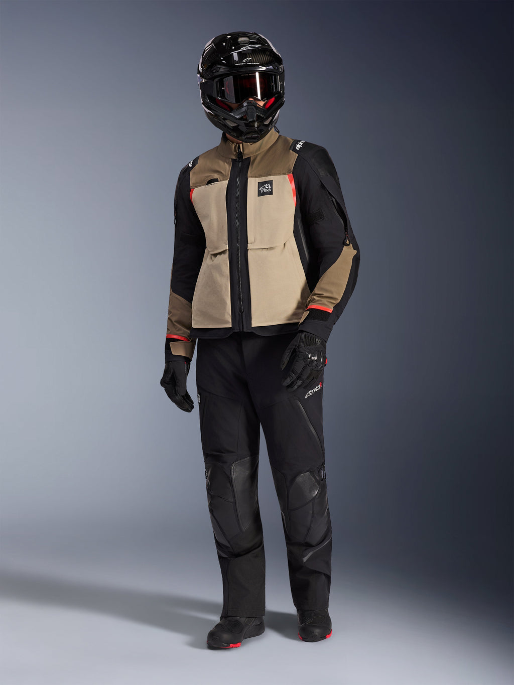 Casaco Alpinestars Andes Pro Drystar® XF em Walnut Aluminum, casaco de motociclismo de aventura usado com calças pretas a condizer, um capacete de aventura preto com óculos e botas pretas, com painéis beges e pretos e detalhes em vermelho.