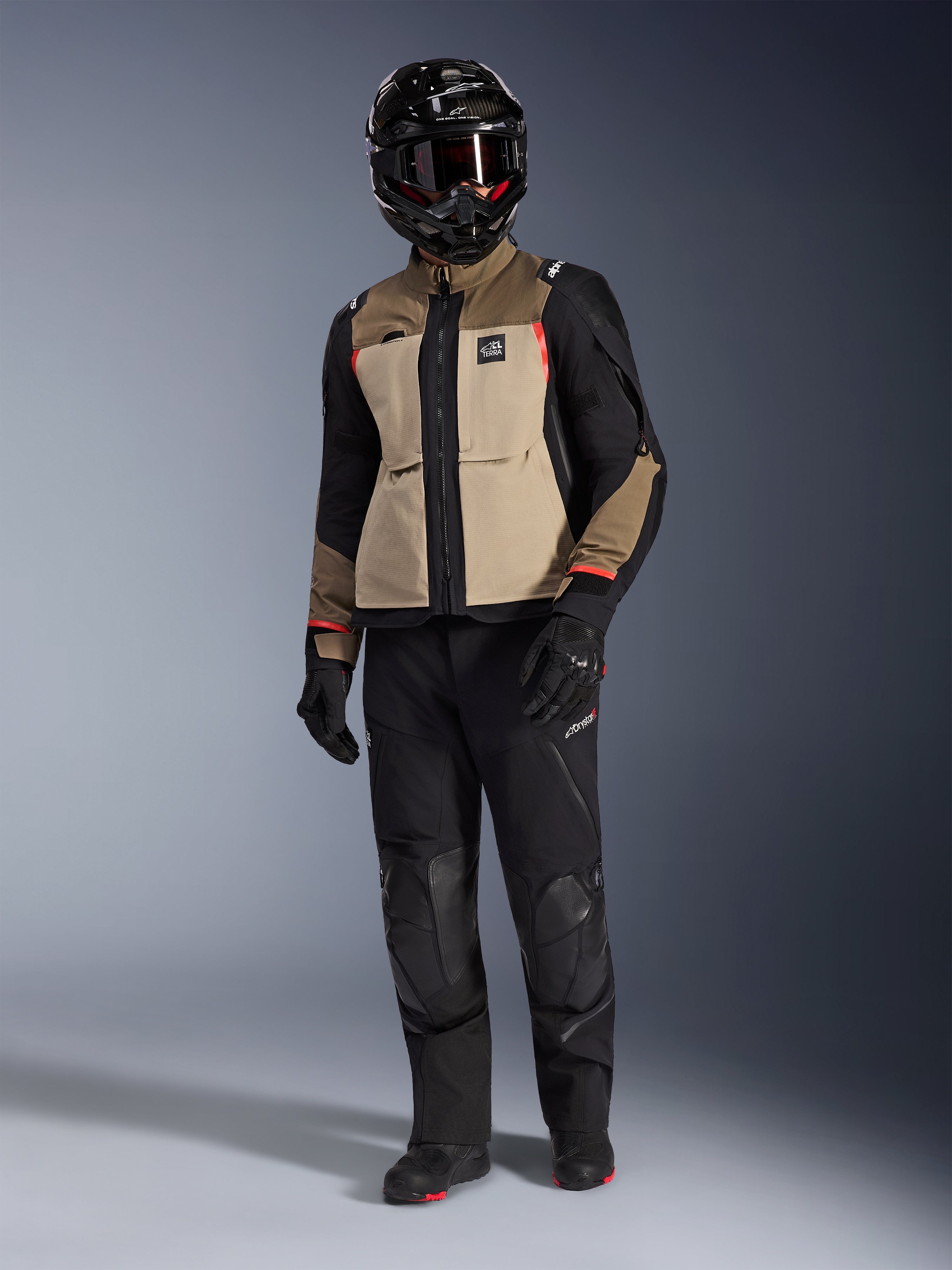 Casaco Alpinestars Andes Pro Drystar® XF em Walnut Aluminum, casaco de motociclismo de aventura usado com calças pretas a condizer, um capacete de aventura preto com óculos e botas pretas, com painéis beges e pretos e detalhes em vermelho.