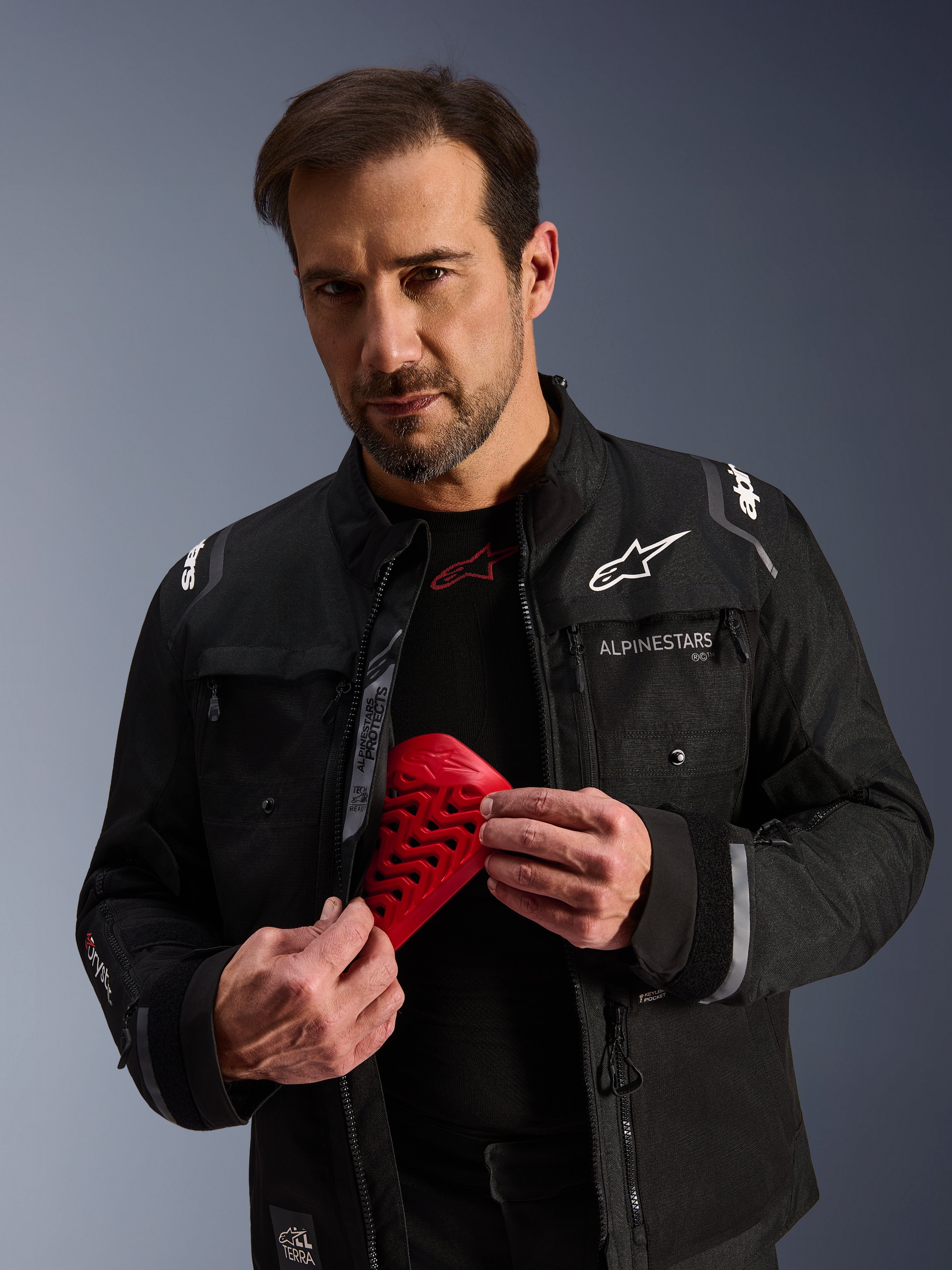Alpinestars Cusco Drystar® Jacket in Black, male model holding a red Nucleon Flex Pro armor piece, adventure touring motorcycle jacket with white branding, technical textile construction featuring ventilation zippers and multiple pockets