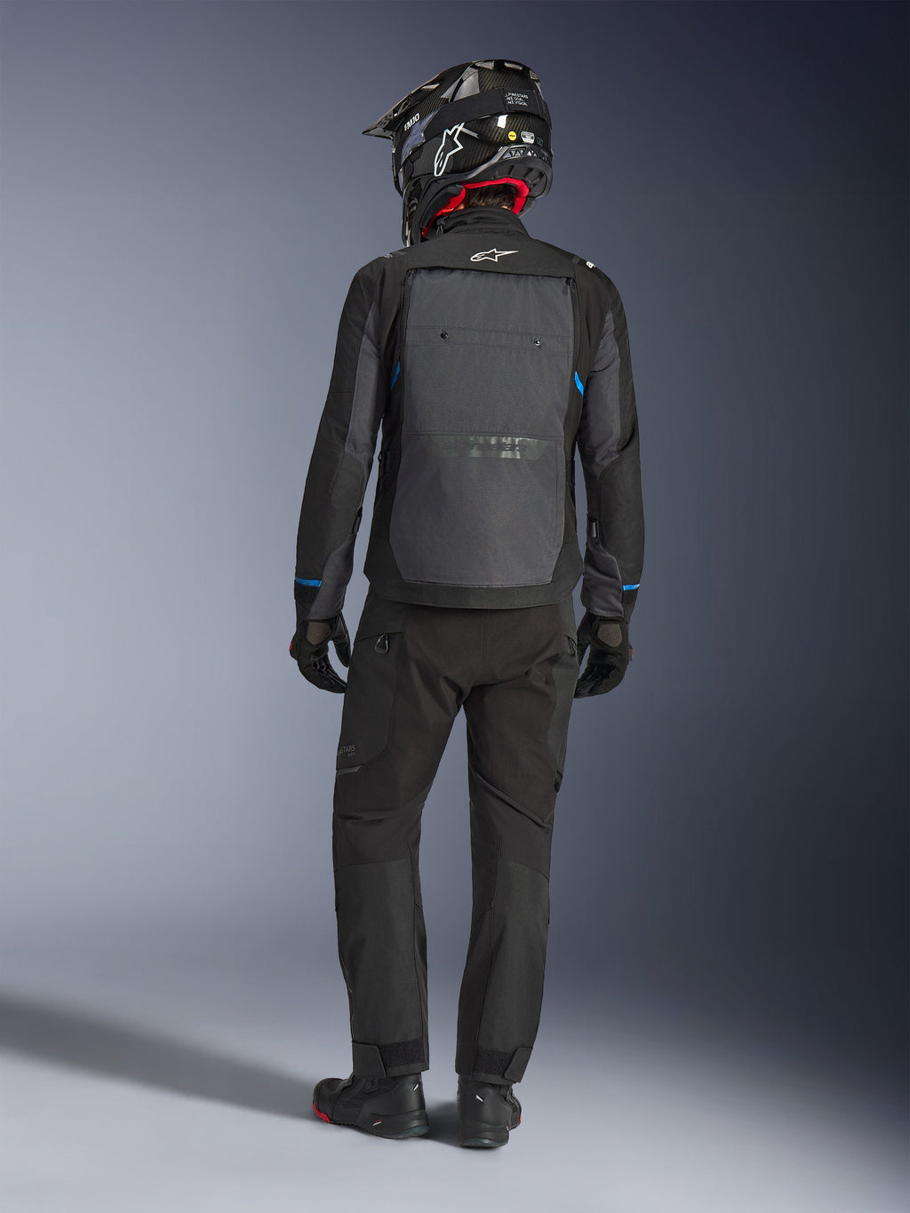 Casaco Alpinestars Cusco Drystar®, preto e cinzento escuro, vista traseira de um condutor com equipamento completo de touring de aventura, grande painel traseiro cinzento com o logótipo Alpinestars, detalhes azuis nas mangas, usado com calças pretas a condizer e capacete.