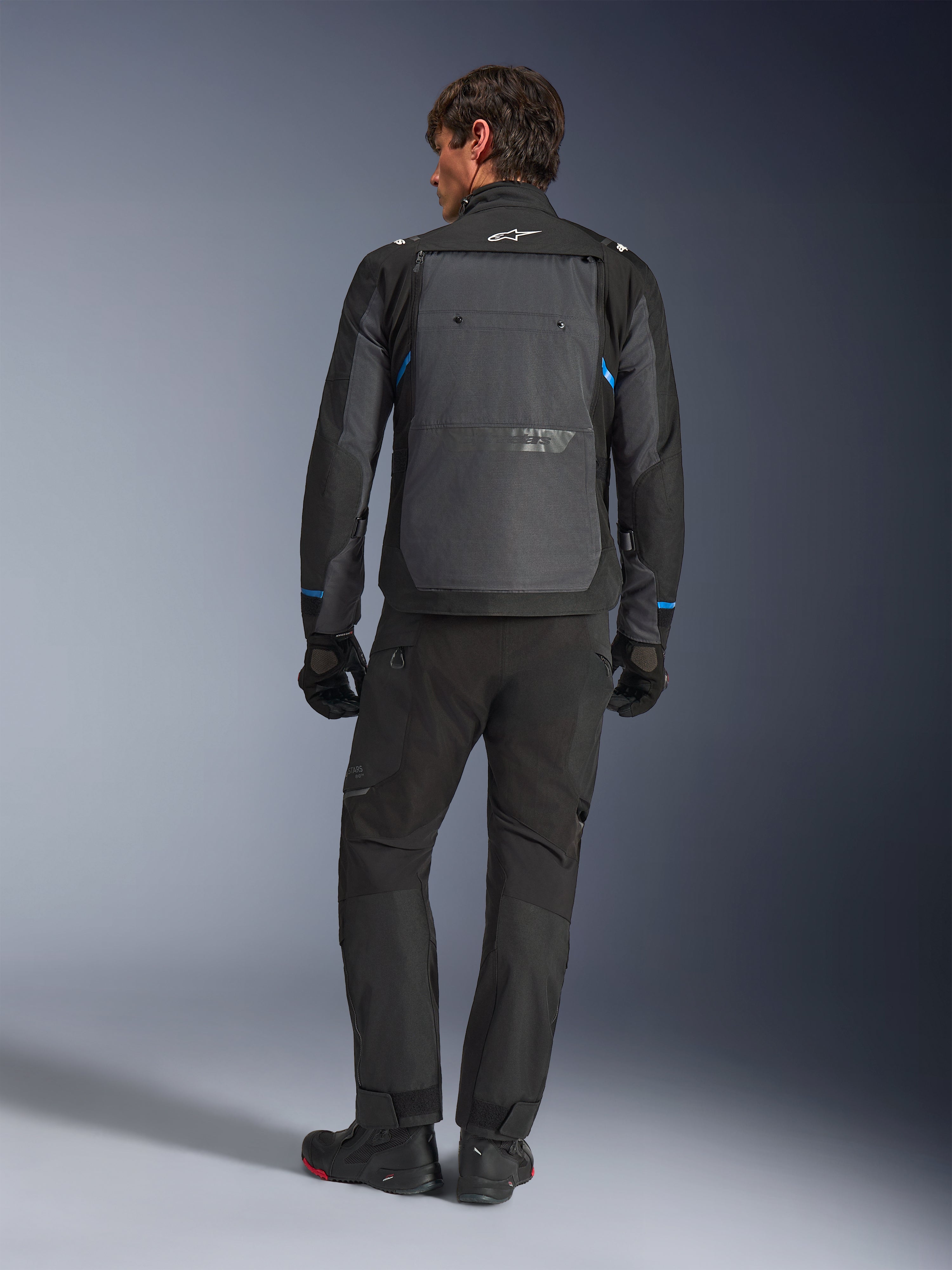 Casaco Alpinestars Cusco Drystar®, preto e cinzento escuro com detalhes azuis, vista traseira, usado com calças de aventura e luvas pretas a condizer, com um grande painel traseiro cinzento e logótipo branco na gola, concebido para estrada e turismo de aventura