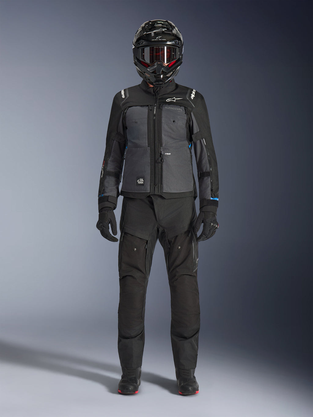 Casaco Alpinestars Cusco Drystar®, casaco de turismo de aventura preto e cinzento escuro, usado com calças pretas a condizer, luvas e um capacete integral, vista frontal de corpo inteiro, vestuário de proteção para motociclismo