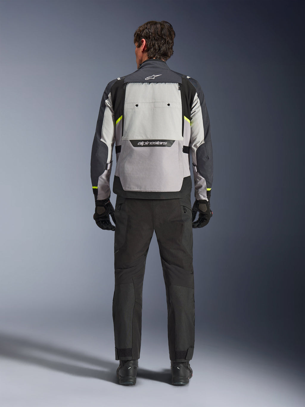 Vista traseira de um motociclista a usar o Casaco Alpinestars Cusco Drystar® em Ice Gray, Mid Gray e Deep Gray, combinado com calças e luvas de motociclismo pretas, destacando o amplo painel de ventilação traseiro e o logótipo Alpinestars.