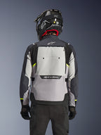 Casaco Alpinestars Cusco Drystar®, vista traseira, Ice Gray, Mid Gray e Deep Gray, com um grande painel de ventilação traseiro e detalhes em amarelo de alta visibilidade, usado com um capacete de adventure preto e calças pretas.