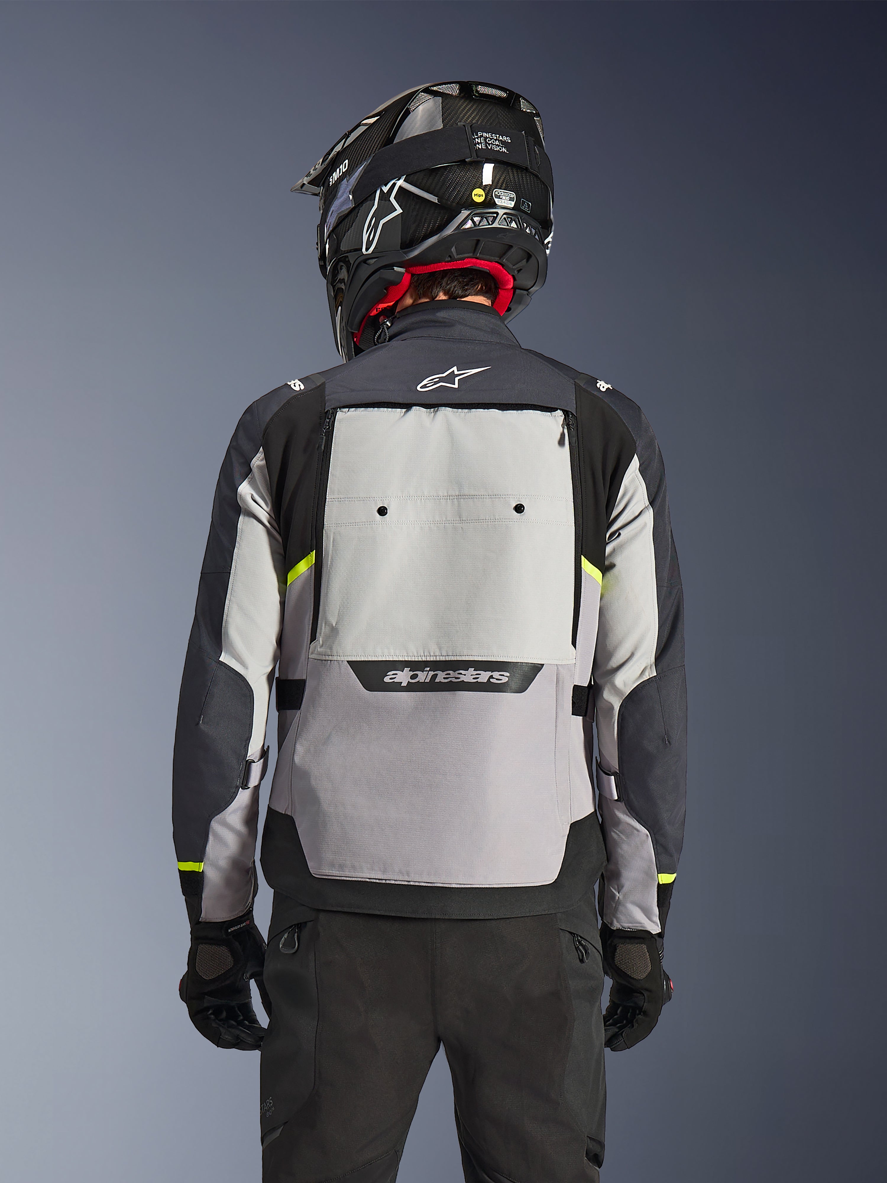 Casaco Alpinestars Cusco Drystar®, vista traseira, Ice Gray, Mid Gray e Deep Gray, com um grande painel de ventilação traseiro e detalhes em amarelo de alta visibilidade, usado com um capacete de adventure preto e calças pretas.