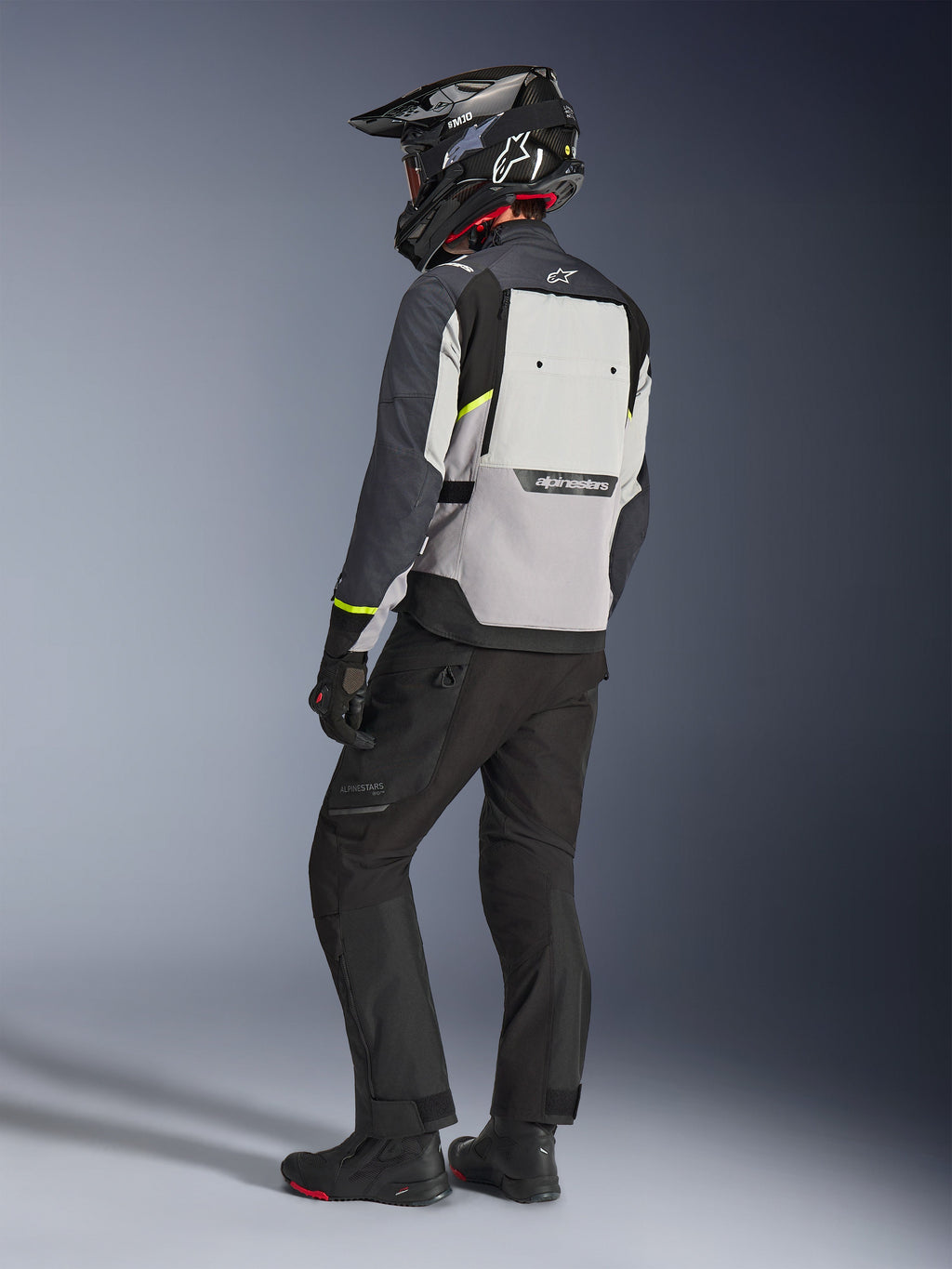 Casaco Alpinestars Cusco Drystar® em Ice Gray, Mid Gray e Deep Gray, vista traseira num motociclista. Apresenta pormenores em amarelo fluorescente, um grande painel de ventilação traseiro com a marca e construção têxtil durável. Usado com calças de motociclismo pretas e um capacete de aventura para um visual touring completo.