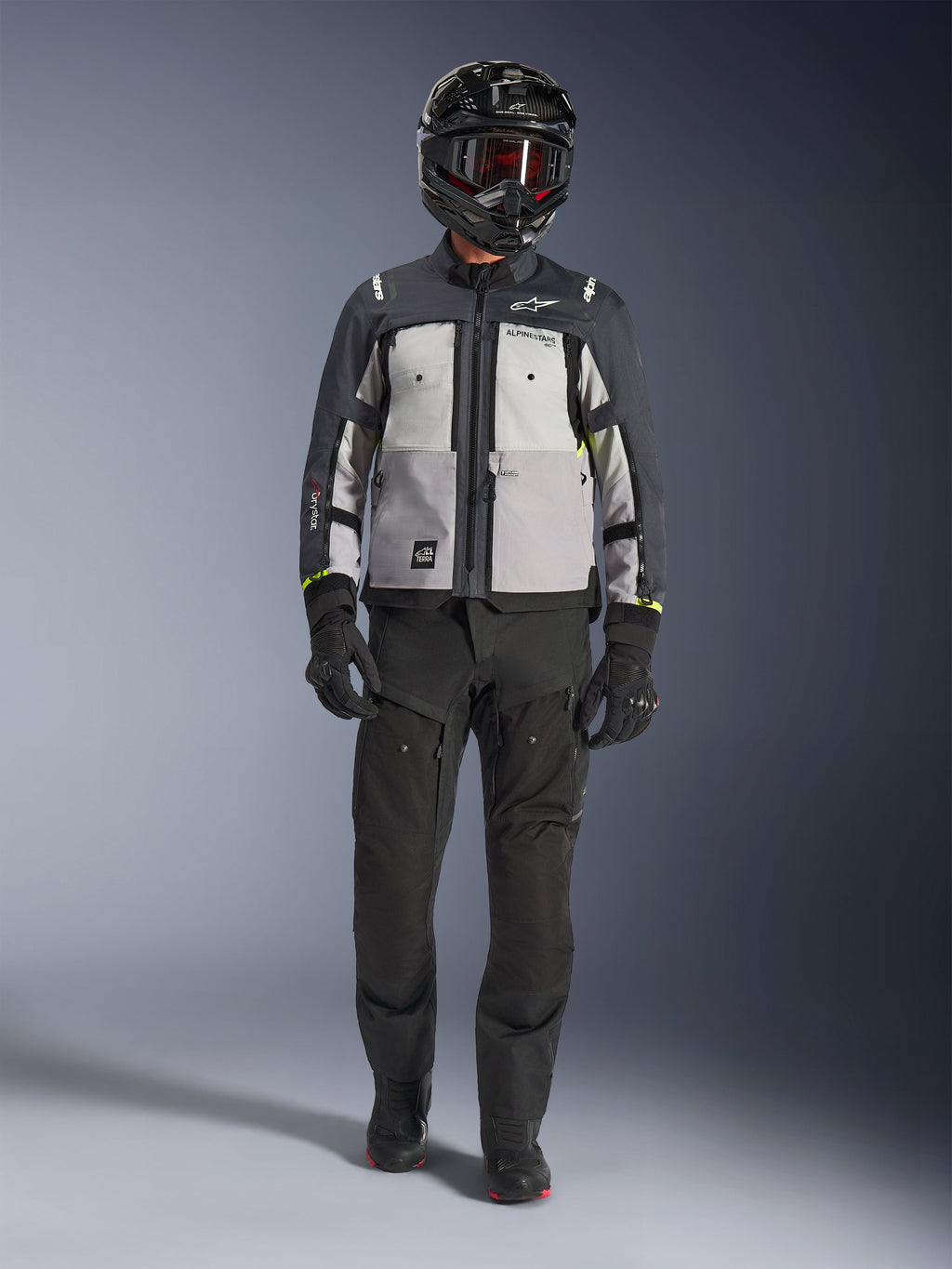 Alpinestars Cusco Drystar® Jacket, cinzento gelo, cinzento médio e cinzento escuro, casaco de motociclismo de aventura usado com calças de condução pretas, luvas pretas e capacete preto com óculos, com painéis cinzentos em blocos de cor e detalhes técnicos para estrada e adventure touring.