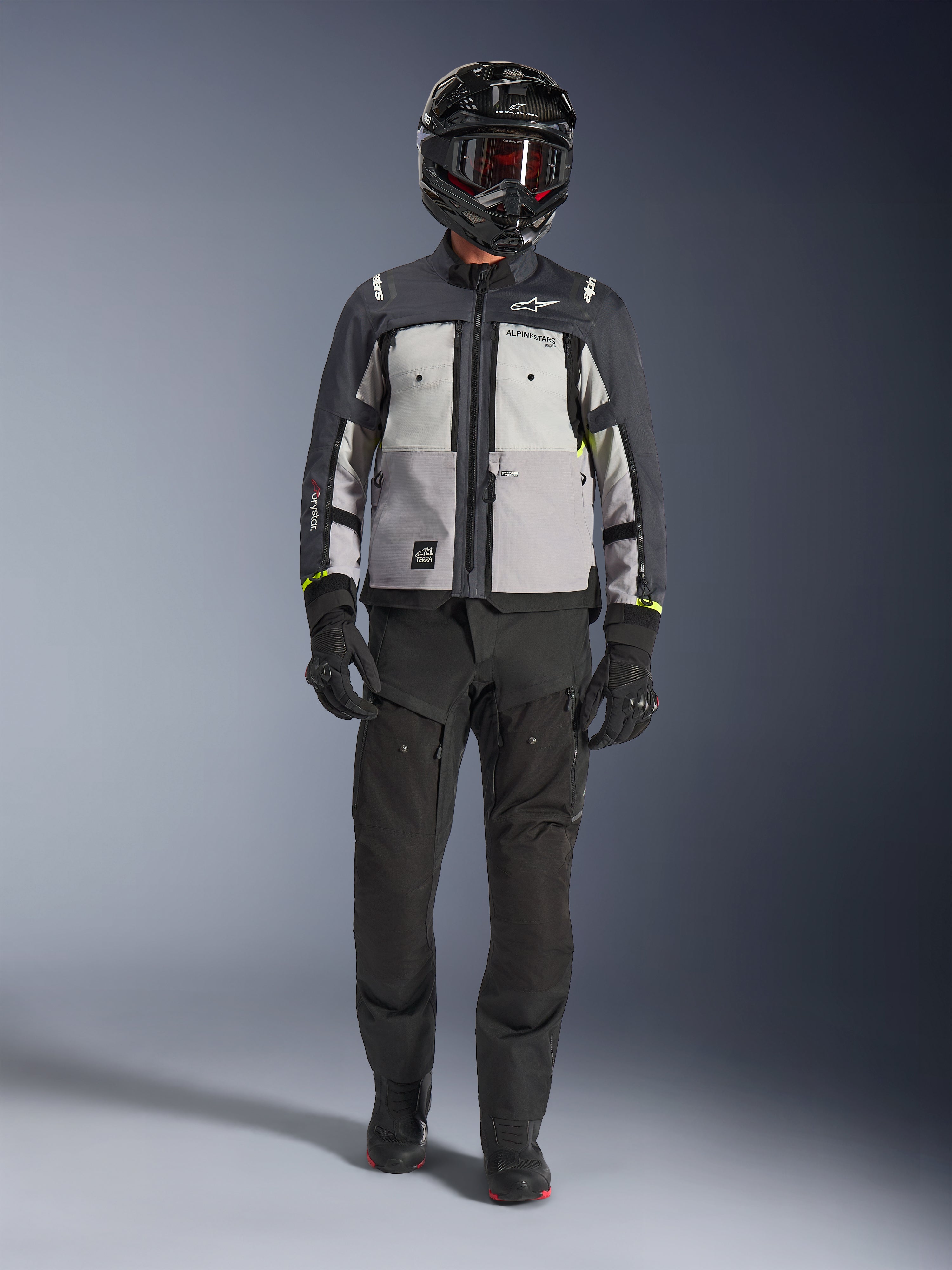 Alpinestars Cusco Drystar® Jacket, cinzento gelo, cinzento médio e cinzento escuro, casaco de motociclismo de aventura usado com calças de condução pretas, luvas pretas e capacete preto com óculos, com painéis cinzentos em blocos de cor e detalhes técnicos para estrada e adventure touring.