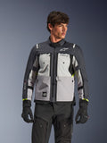 Casaco Alpinestars Cusco Drystar®, casaco de motociclismo adventure touring, cinzento gelo, cinzento médio e cinzento escuro, usado por um modelo com calças e luvas pretas, design de tecido técnico multi-painéis, bolsos no peito, marca 'Drystar' na manga e detalhes subtis em amarelo néon nos punhos