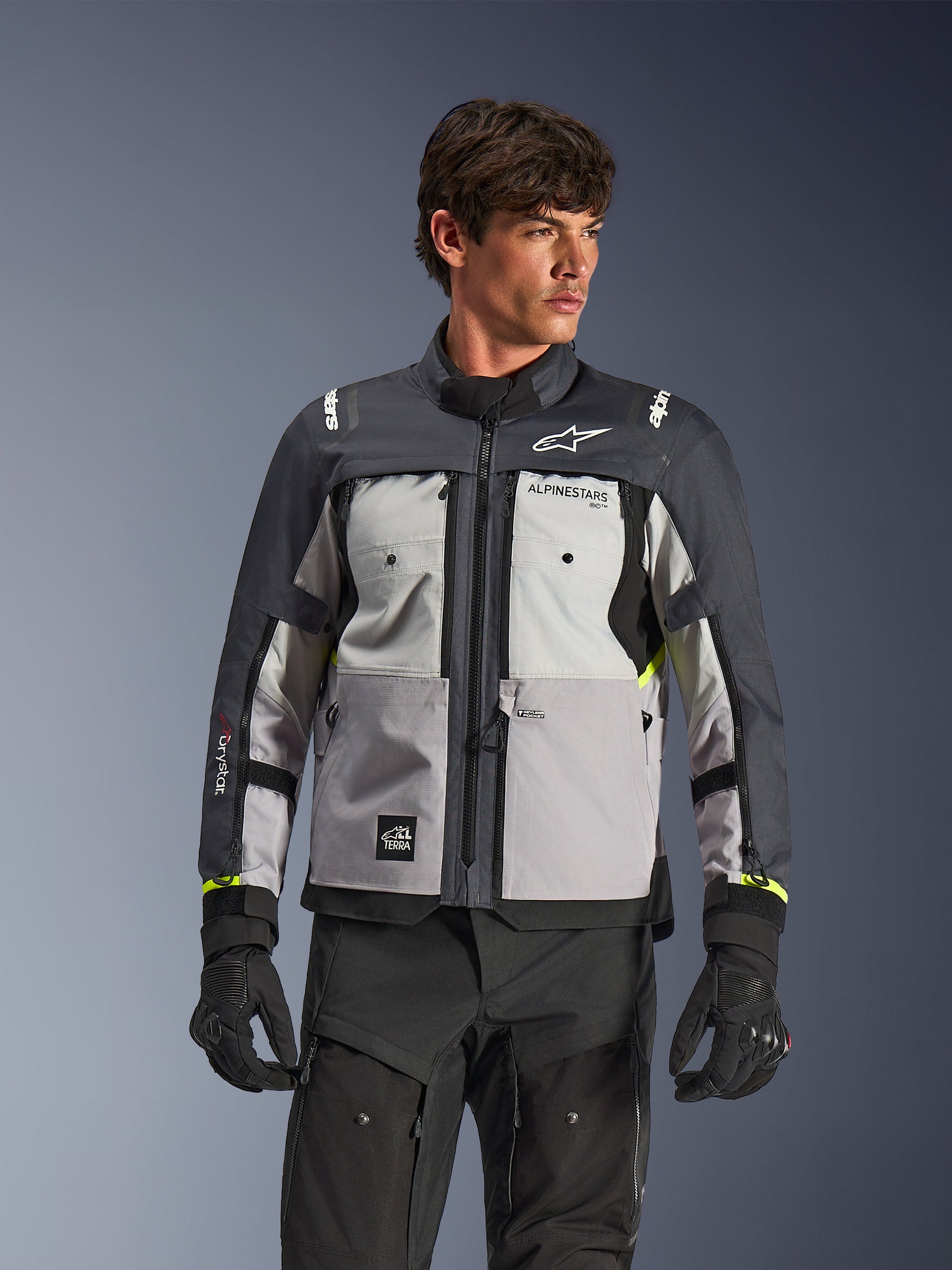 Alpinestars Cusco Drystar® Jacket, Adventure Touring Motorcycle Jacket, Ice Gray, Mid Gray, and Deep Gray, worn by a model with black pants and gloves, featuring a multi-paneled technical textile design, chest pockets, 'Drystar' branding on the sleeve, and subtle neon yellow accents at the cuffs.
