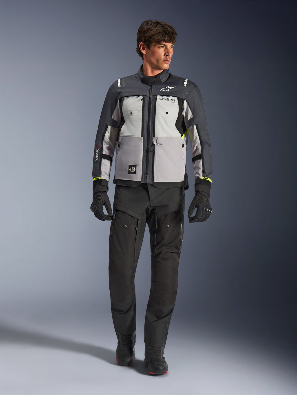 Casaco Alpinestars Cusco Drystar® em cinzento gelo, cinzento médio e cinzento escuro, usado com calças e luvas de motociclismo de aventura pretas, casaco de touring de aventura em têxtil com painéis em vários tons de cinzento e detalhes em amarelo néon, fotografia de estúdio de modelo masculino.