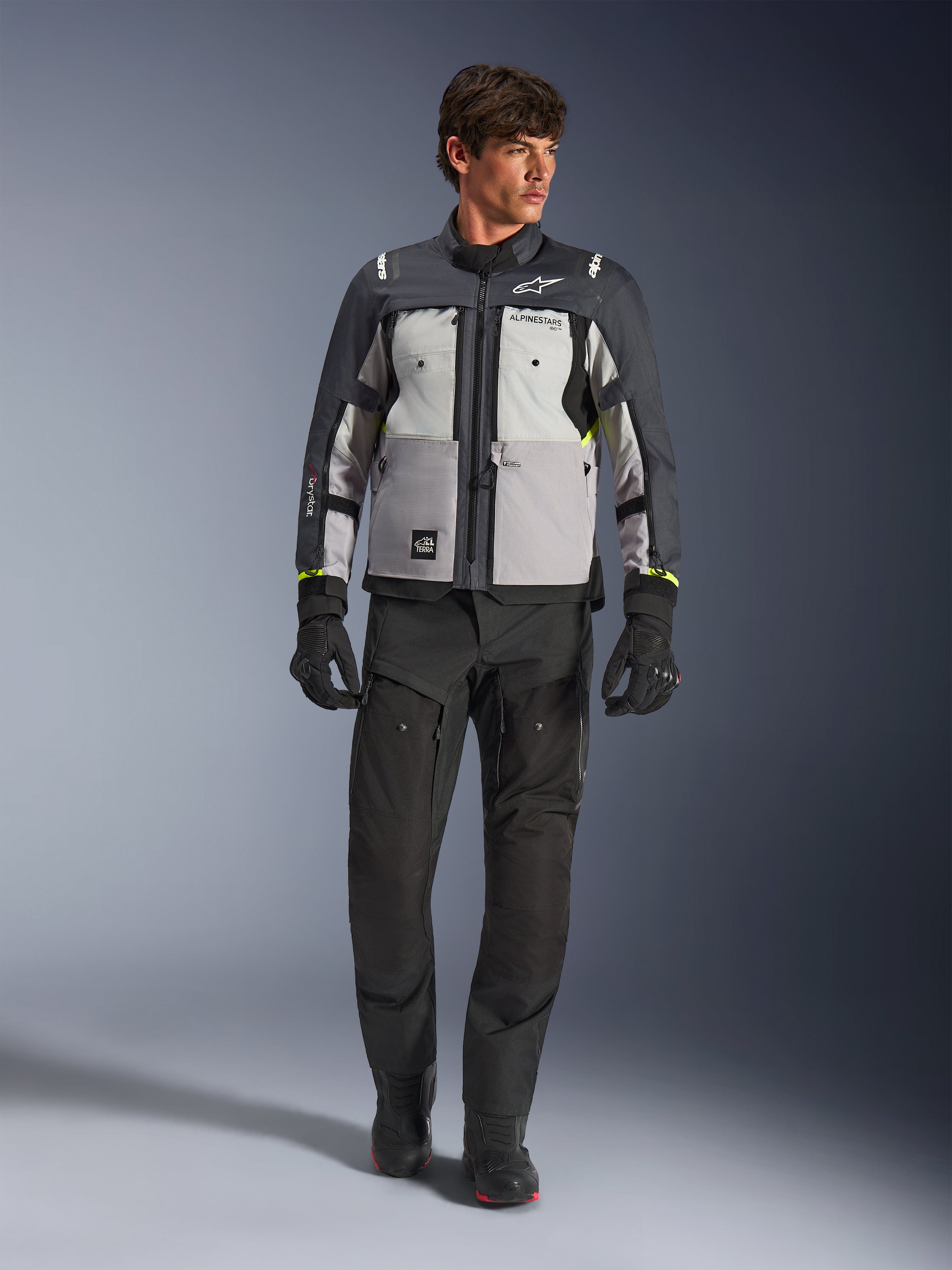 Casaco Alpinestars Cusco Drystar® em cinzento gelo, cinzento médio e cinzento escuro, usado com calças e luvas de motociclismo de aventura pretas, casaco de touring de aventura em têxtil com painéis em vários tons de cinzento e detalhes em amarelo néon, fotografia de estúdio de modelo masculino.
