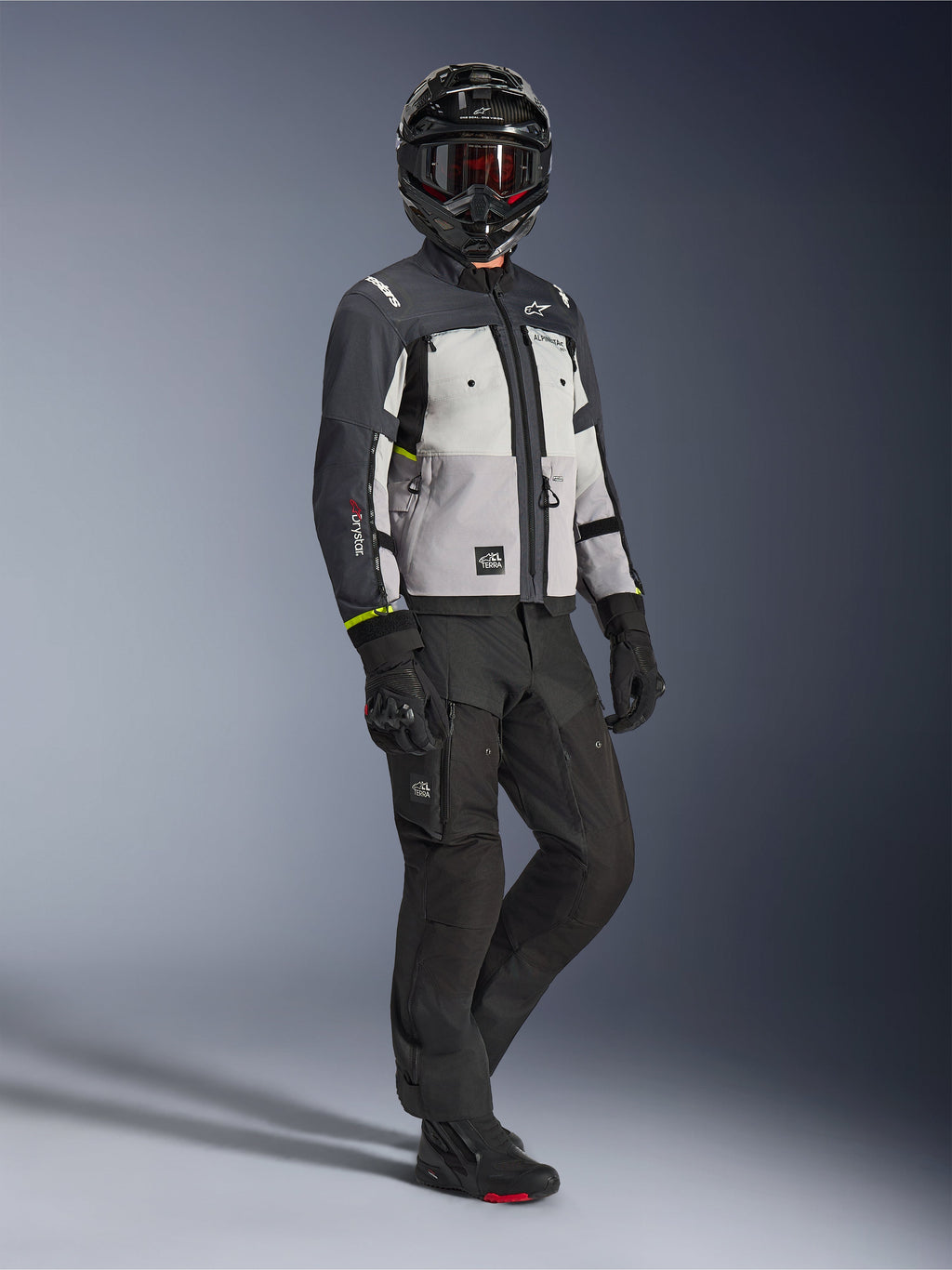 Casaco Alpinestars Cusco Drystar® em Ice Gray, Mid Gray e Deep Gray, fotografia de corpo inteiro de um motociclista com equipamento completo de aventura, incluindo capacete preto com óculos vermelhos, calças pretas e luvas, casaco de motociclismo têxtil com painéis de ventilação direta e membrana impermeável