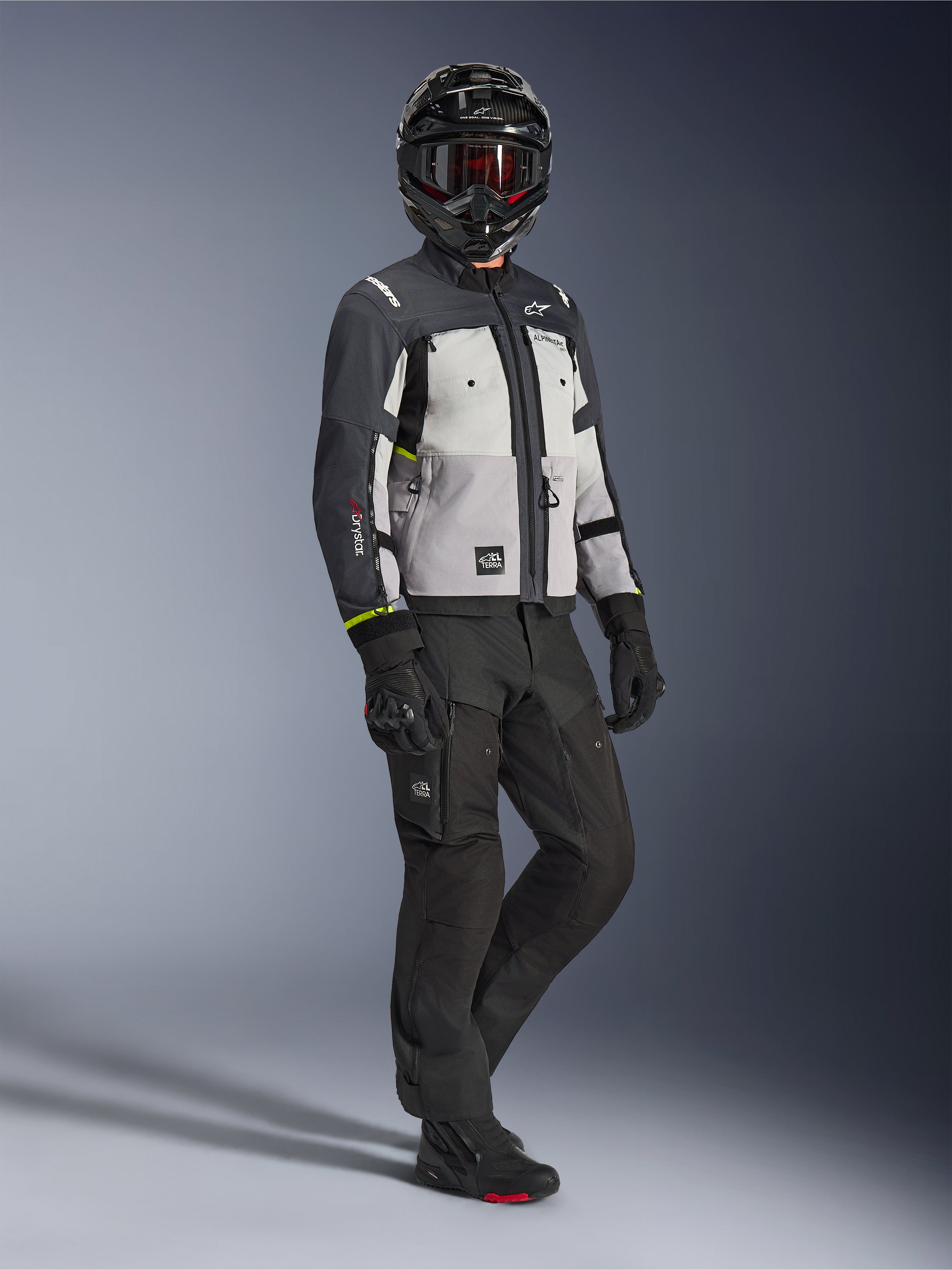 Casaco Alpinestars Cusco Drystar® em Ice Gray, Mid Gray e Deep Gray, fotografia de corpo inteiro de um motociclista com equipamento completo de aventura, incluindo capacete preto com óculos vermelhos, calças pretas e luvas, casaco de motociclismo têxtil com painéis de ventilação direta e membrana impermeável