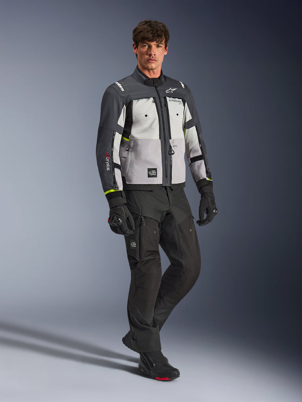 Casaco Alpinestars Cusco Drystar®, casaco de motociclismo adventure, cinzento gelo, cinzento médio e cinzento escuro, usado por um modelo com calças e luvas pretas, painéis cinzentos multi-tons com construção têxtil durável, com aberturas de ventilação com fecho e logótipos Alpinestars, fotografia de estúdio profissional para equipamento de estrada e aventura.