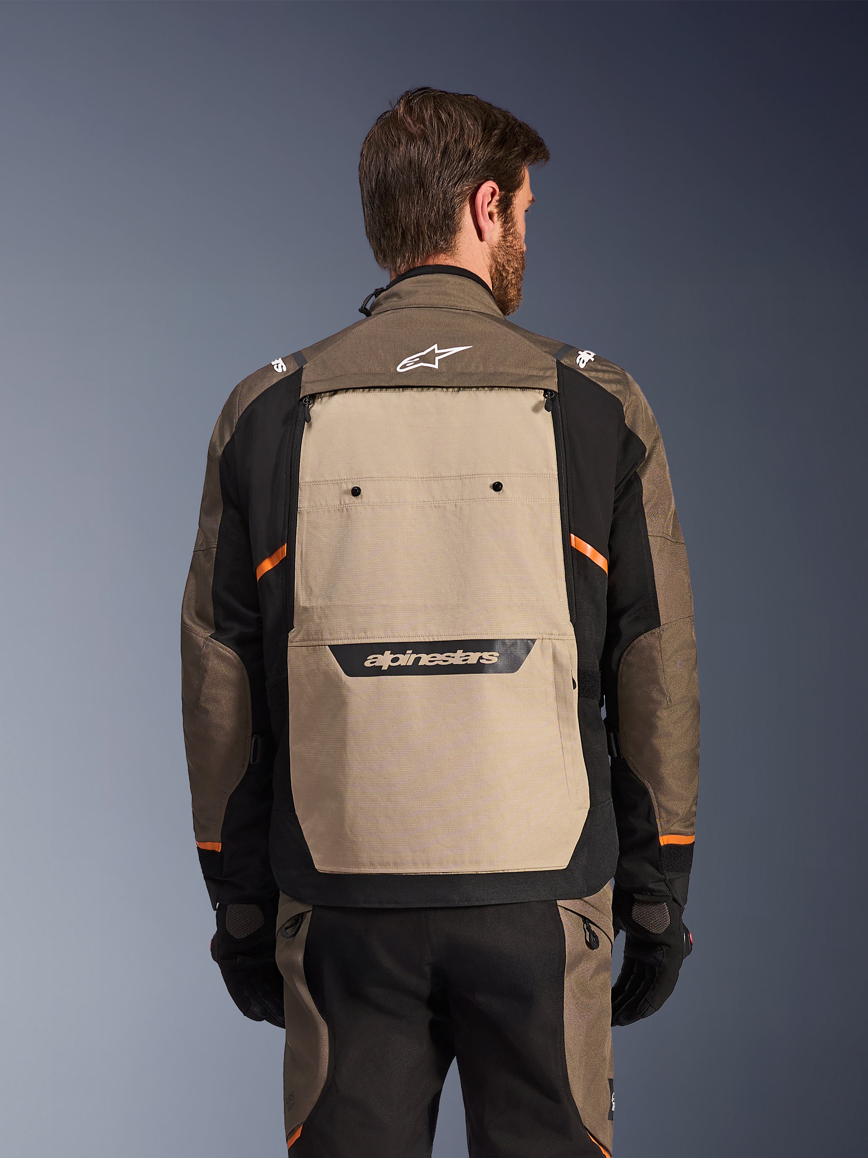 Casaco Alpinestars Cusco Drystar®, Walnut Aluminum Black, vista traseira, com um grande painel traseiro bege e logótipo preto, detalhes laranja, usado com calças e luvas a condizer, concebido para adventure touring