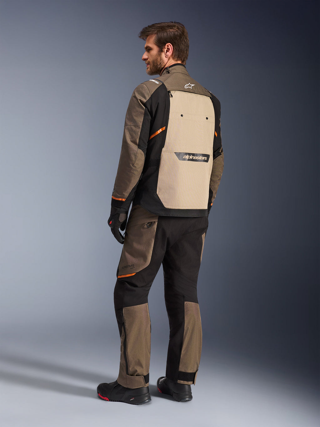 Casaco Alpinestars Cusco Drystar® em Walnut Aluminum Black, vista traseira mostrando o painel traseiro bege e detalhes em laranja, usado com calças de motociclismo a condizer, luvas e botas pretas, equipamento profissional de touring de aventura