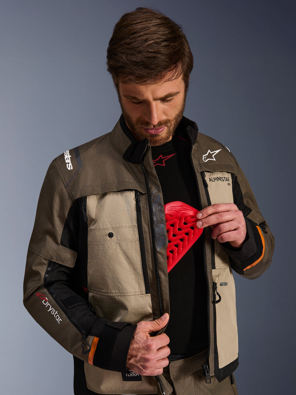 Casaco Alpinestars Cusco Drystar®, Walnut Aluminum Black, vestido por um homem a demonstrar o bolso para o protetor de peito com uma inserção de proteção vermelha, casaco de motociclismo têxtil com aberturas de ventilação com fecho e branding para adventure touring.