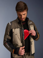 Casaco Alpinestars Cusco Drystar®, Walnut Aluminum Black, vestido por um homem a demonstrar o bolso para o protetor de peito com uma inserção de proteção vermelha, casaco de motociclismo têxtil com aberturas de ventilação com fecho e branding para adventure touring.