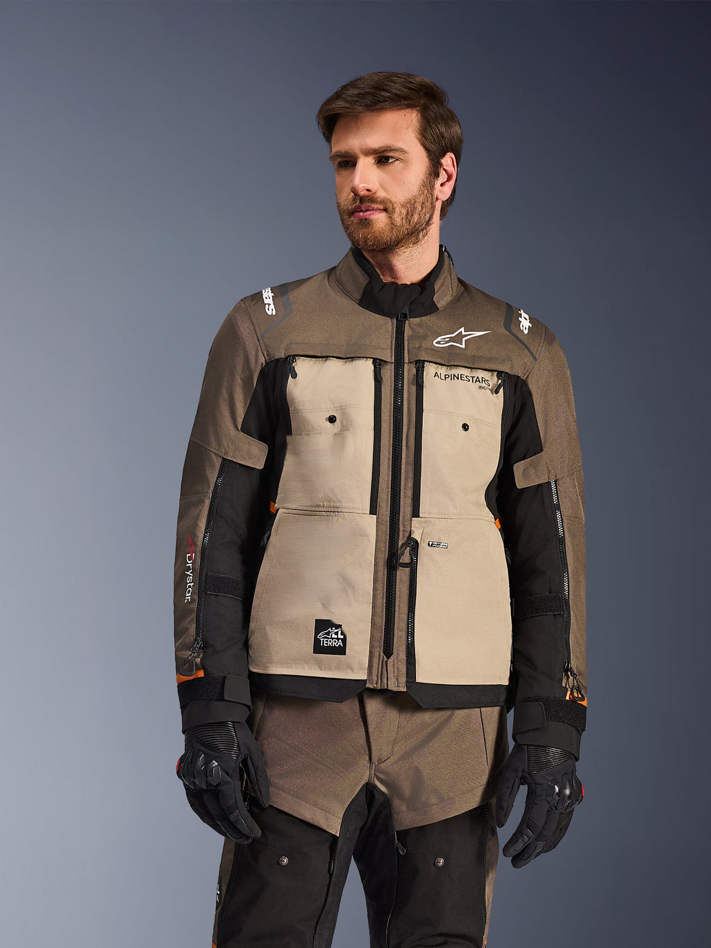Casaco Alpinestars Cusco Drystar®, casaco de aventura para motociclismo, na cor nogueira, alumínio e preto, usado por um modelo com luvas pretas, com painéis beges no peito e mangas pretas com a marca Alpinestars e emblema All Terra, equipamento de touring protetor para todas as condições meteorológicas