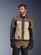 Casaco Alpinestars Cusco Drystar®, casaco de aventura para motociclismo, na cor nogueira, alumínio e preto, usado por um modelo com luvas pretas, com painéis beges no peito e mangas pretas com a marca Alpinestars e emblema All Terra, equipamento de touring protetor para todas as condições meteorológicas