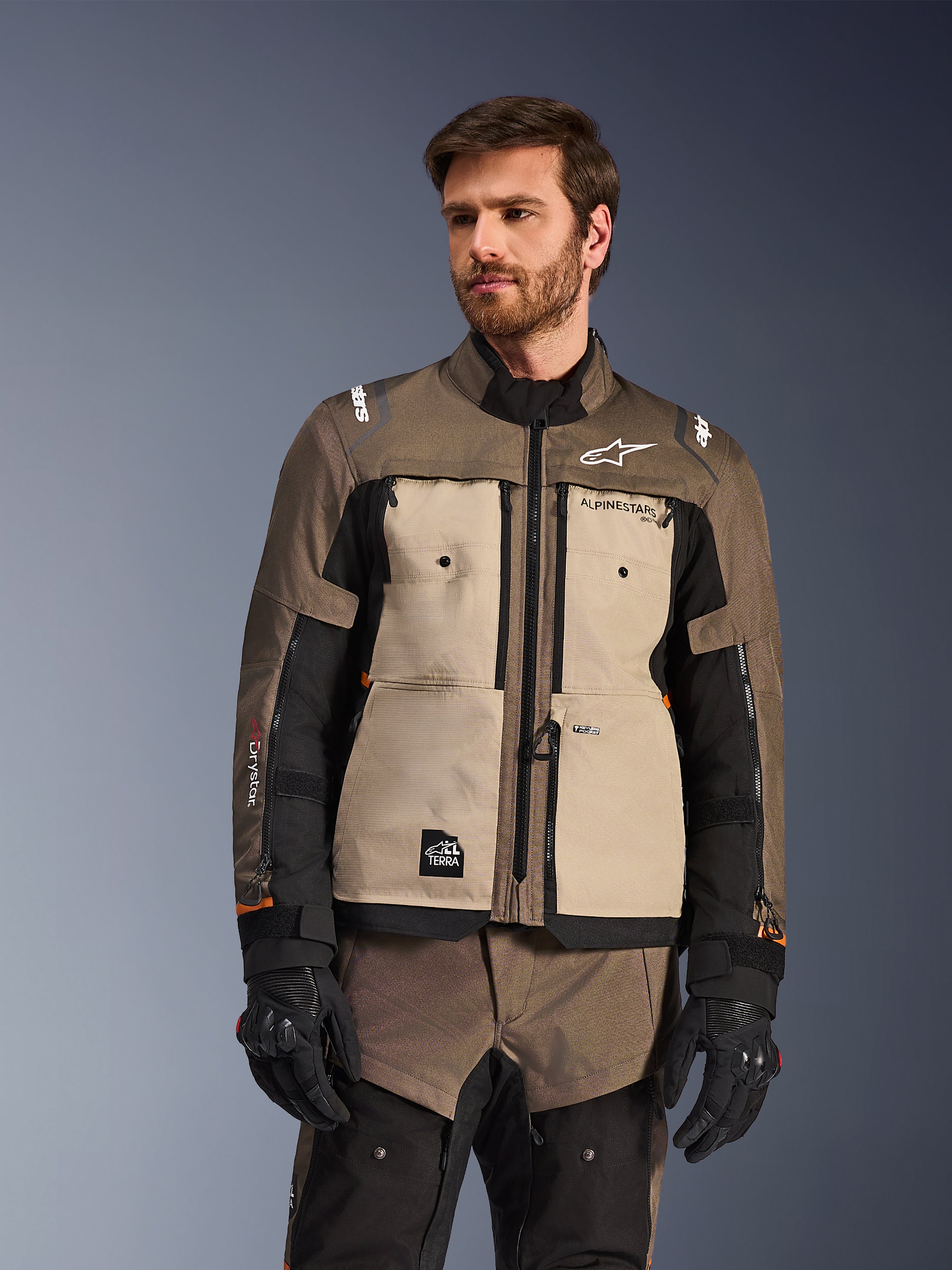 Casaco Alpinestars Cusco Drystar®, casaco de aventura para motociclismo, na cor nogueira, alumínio e preto, usado por um modelo com luvas pretas, com painéis beges no peito e mangas pretas com a marca Alpinestars e emblema All Terra, equipamento de touring protetor para todas as condições meteorológicas