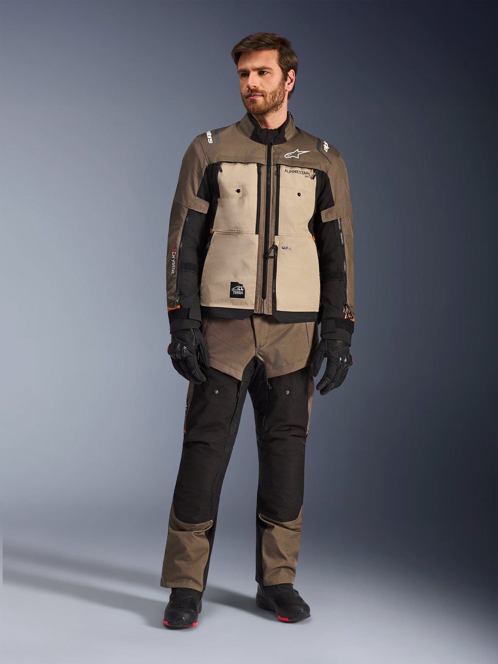 Casaco Alpinestars Cusco Drystar® em Walnut Aluminum Black, usado por um motociclista com calças de adventure touring a condizer, luvas e botas pretas, com painéis no peito em bege, mangas pretas com detalhes subtis em cor de laranja e fechos de ventilação funcionais para condução de motociclo em estrada.