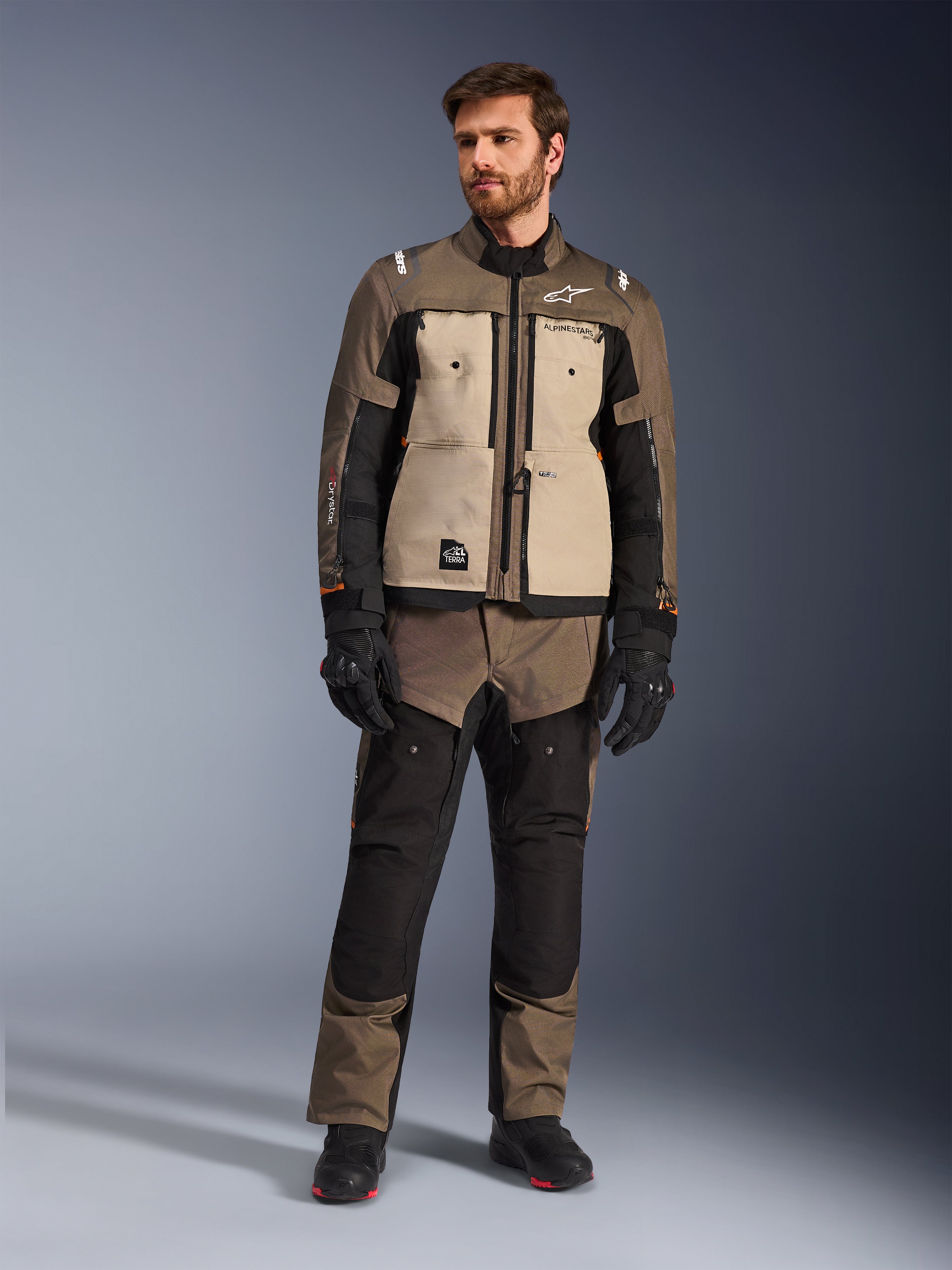 Casaco Alpinestars Cusco Drystar® em Walnut Aluminum Black, usado por um motociclista com calças de adventure touring a condizer, luvas e botas pretas, com painéis no peito em bege, mangas pretas com detalhes subtis em cor de laranja e fechos de ventilação funcionais para condução de motociclo em estrada.