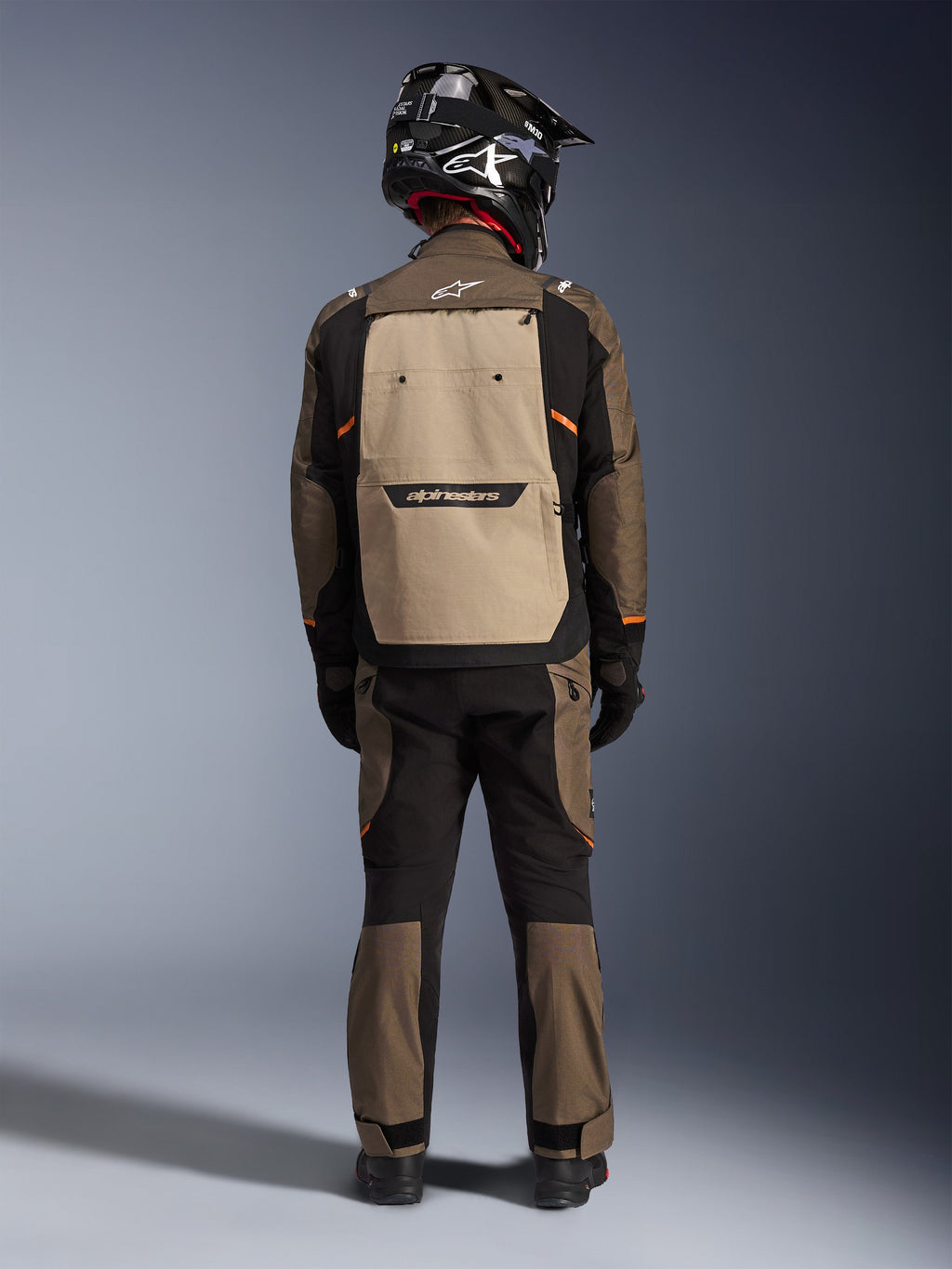 Motociclista visto de costas a usar o Casaco Alpinestars Cusco Drystar® em Walnut Aluminum Black, com um grande bolso traseiro cor de alumínio e detalhes em laranja, calças de aventura a condizer, capacete de off-road preto e luvas, equipamento de motociclismo em têxtil resistente para touring