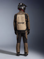 Motociclista visto de costas a usar o Casaco Alpinestars Cusco Drystar® em Walnut Aluminum Black, com um grande bolso traseiro cor de alumínio e detalhes em laranja, calças de aventura a condizer, capacete de off-road preto e luvas, equipamento de motociclismo em têxtil resistente para touring
