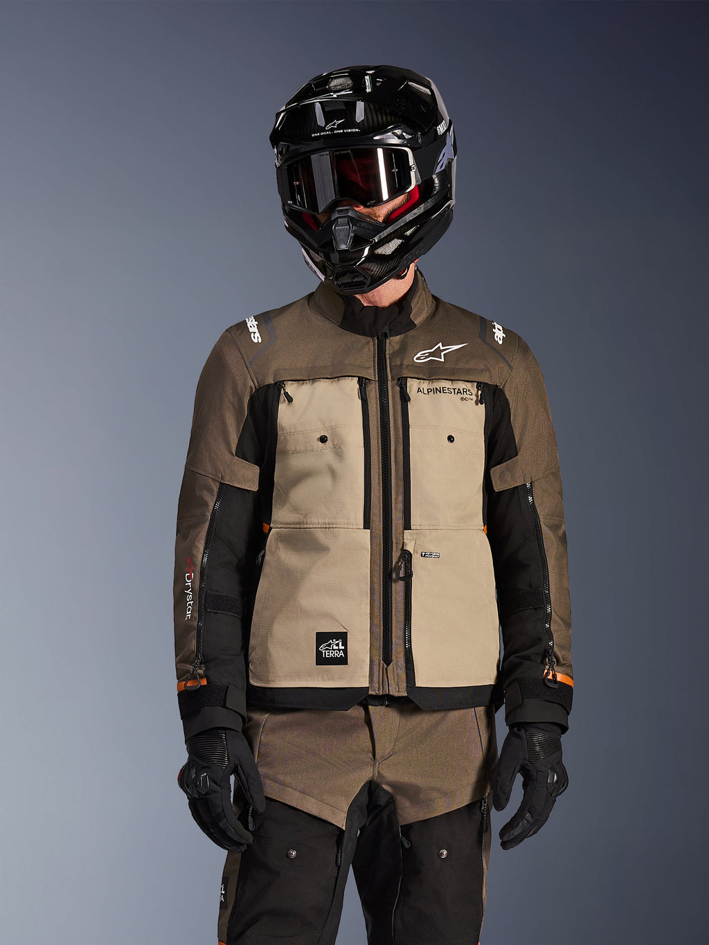 Motociclista a usar o Casaco Cusco Drystar da Alpinestars em Walnut Aluminum Black, equipamento de motociclismo de aventura e touring, capacete preto com óculos e luvas pretas, vista frontal exibindo a construção têxtil técnica, bolsos e logótipos da marca.