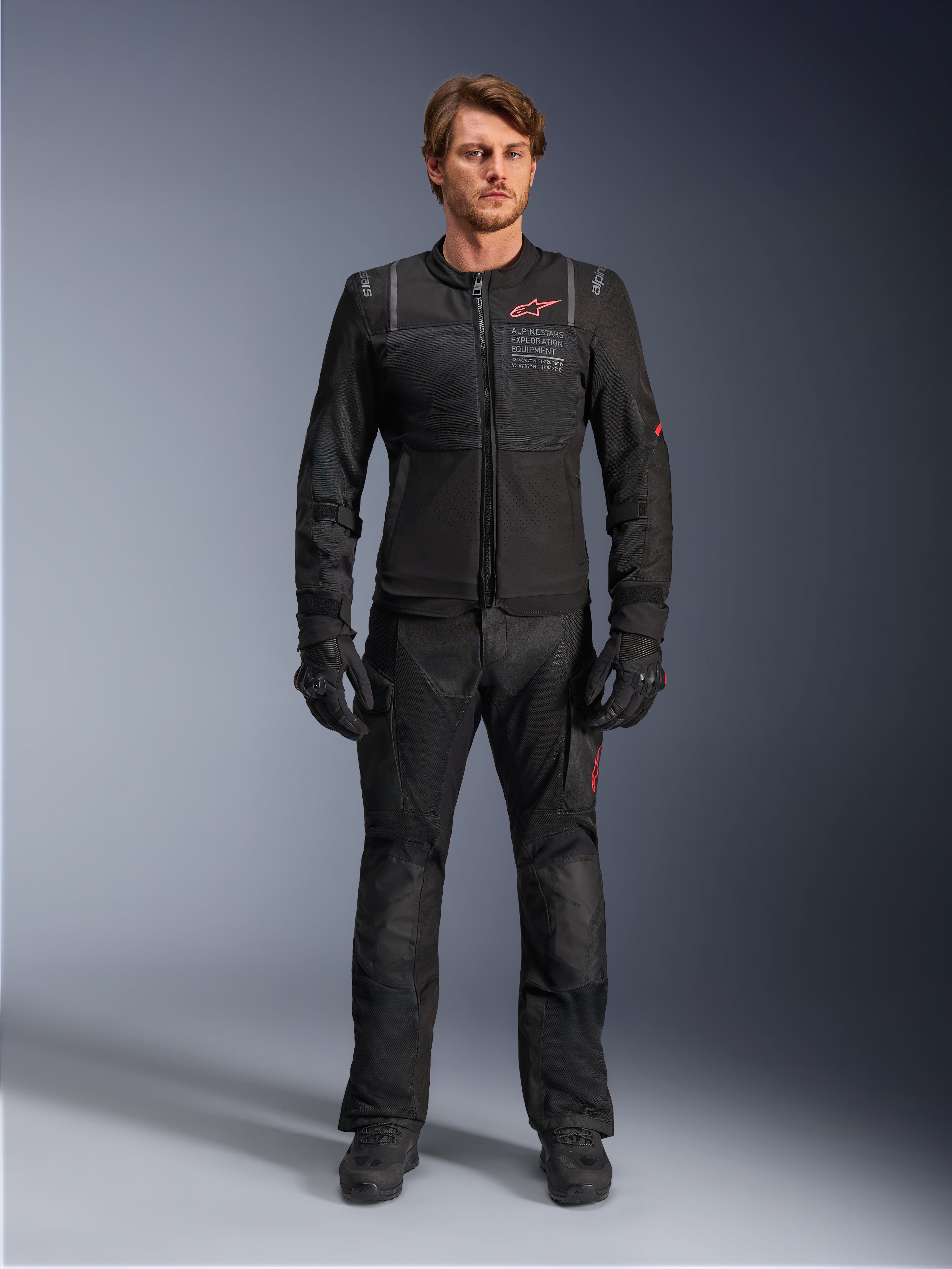 Male model wearing Alpinestars ST-2 Air Jacket, Black motorcycle jacket with mesh panels and red logo, 'Alpinestars Exploration Equipment' text on chest, paired with black technical riding pants and gloves, modern adventure touring gear