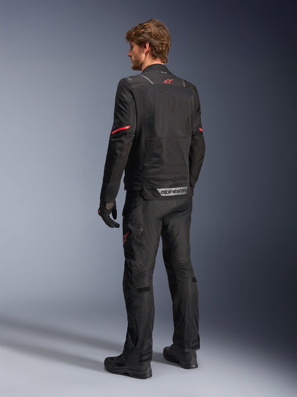 Alpinestars ST-2 Air Jacket, Black motorcycle jacket, rear view on model featuring a red star logo on the upper back and reflective Alpinestars branding at the waist, paired with matching black technical riding pants and gloves.
