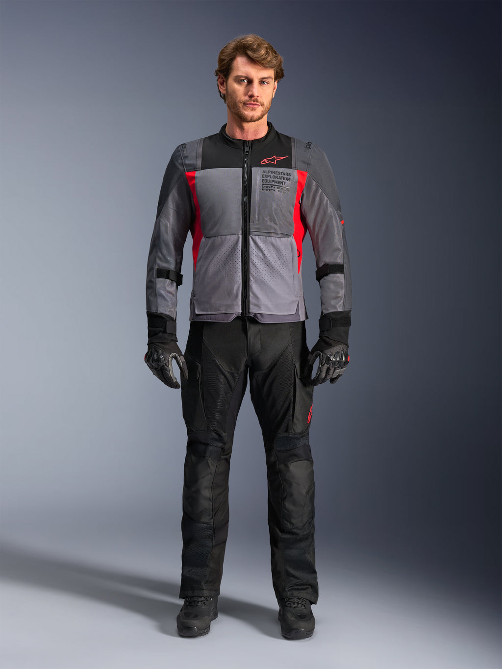 Alpinestars ST-2 Air Jacket in Deep Gray and Bright Red, worn by a male model with black motorcycle pants and gloves, featuring large mesh panels for airflow and modern exploration equipment branding