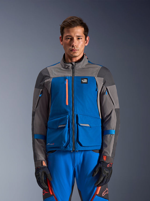 Alpinestars Maxdura Drystar®XF Laminated Armored Shell, casaco de motociclismo de aventura, azul royal e cinzento escuro, usado com calças azuis a condizer e luvas pretas com detalhes cor de laranja, apresenta design com vários bolsos, fecho de correr no peito em cor de laranja e marca 'All Terra', aspeto técnico robusto para condução de aventura