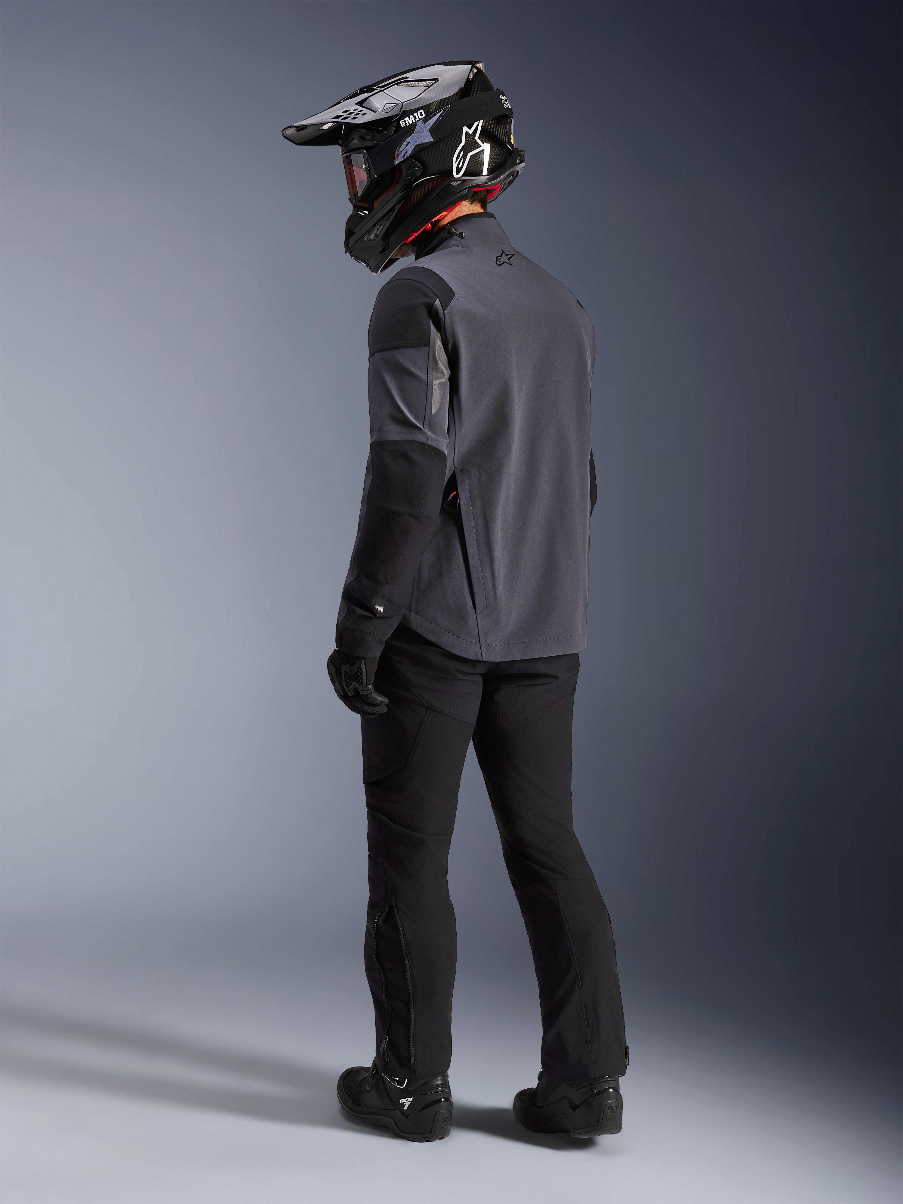 Adventure rider wearing the Maxdura Drystar®XF Laminated Armored Shell in Deep Gray Black, back view. Complete gear setup including black motorcycle pants, a black motocross helmet with Alpinestars branding, and black off-road boots. Technical all-weather riding kit for dual-sport performance.