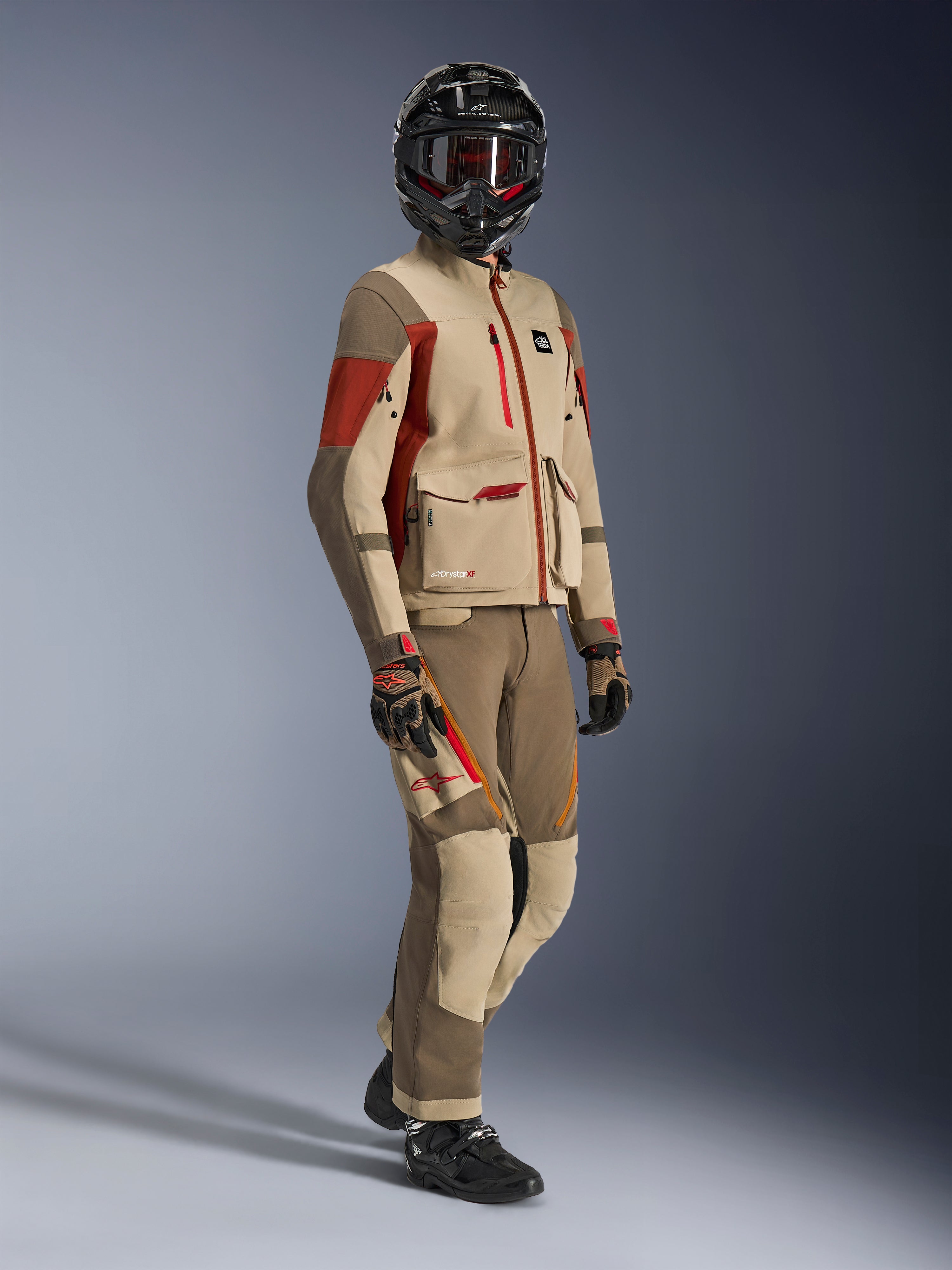 Adventure rider wearing Maxdura Drystar®XF Laminated Armored Shell, Aluminum Walnut and Rust Brown jacket with matching walnut and tan pants, black carbon fiber helmet, and black motorcycle boots, technical touring gear for adventure motorcycling