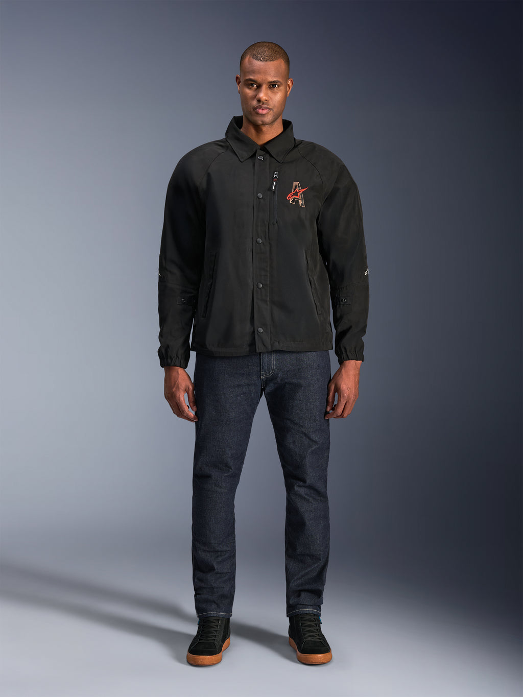 Alpinestars Revved WR Jacket in Black, worn by a model with dark blue jeans and black sneakers, front view, featuring snap button closure and a 3D embroidered logo on the chest, urban contemporary motorcycle gear.